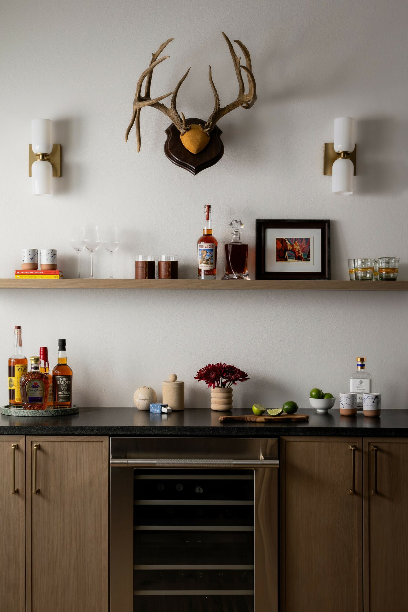 Kitchen with a mounted deer antler display, two wall sconces, and a shelf with bottles, framed photo, and decor items.