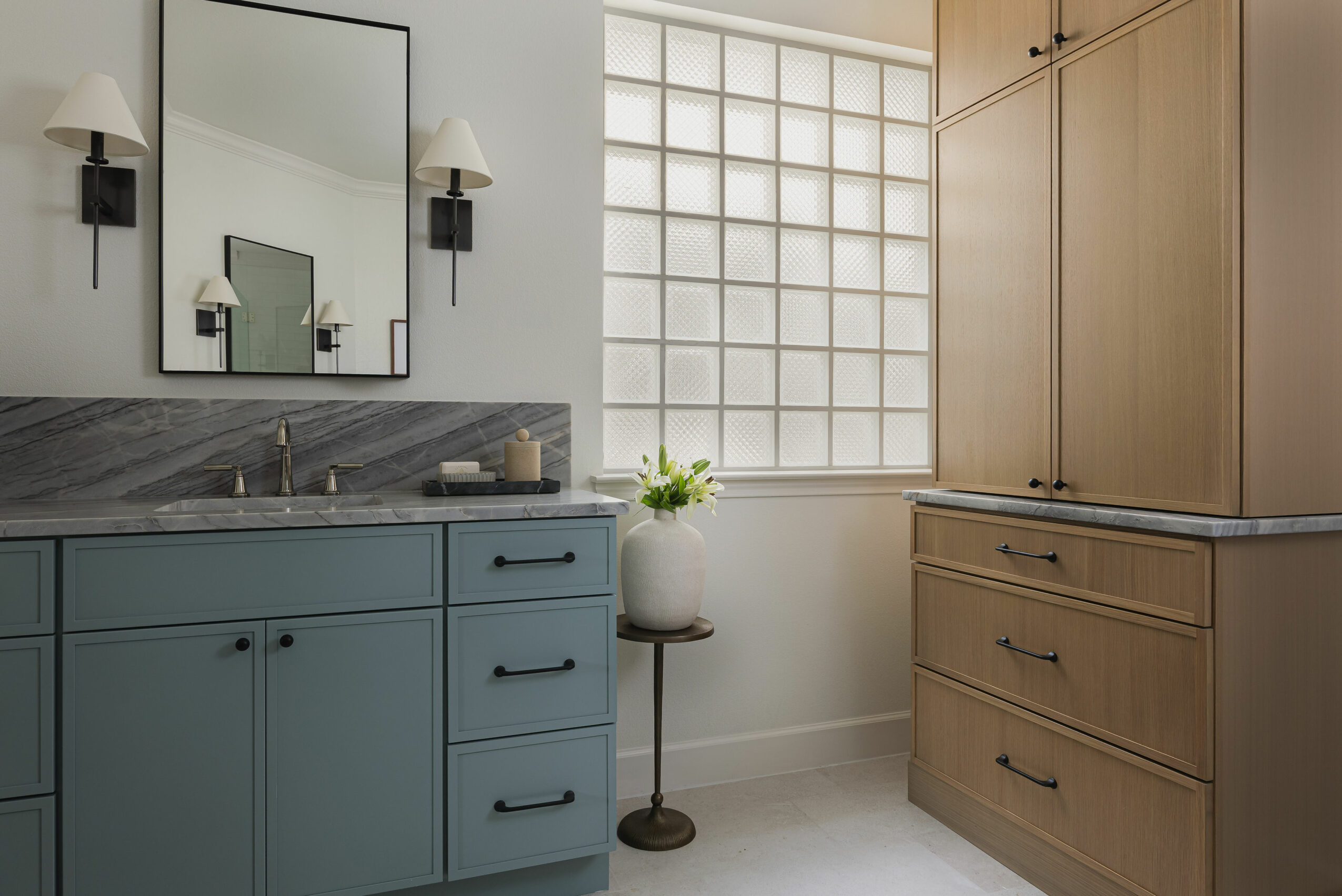 Bathroom with blue cabinet, mirror, wall sconces, glass block window, and wooden storage cabinet, with a small table and plant.