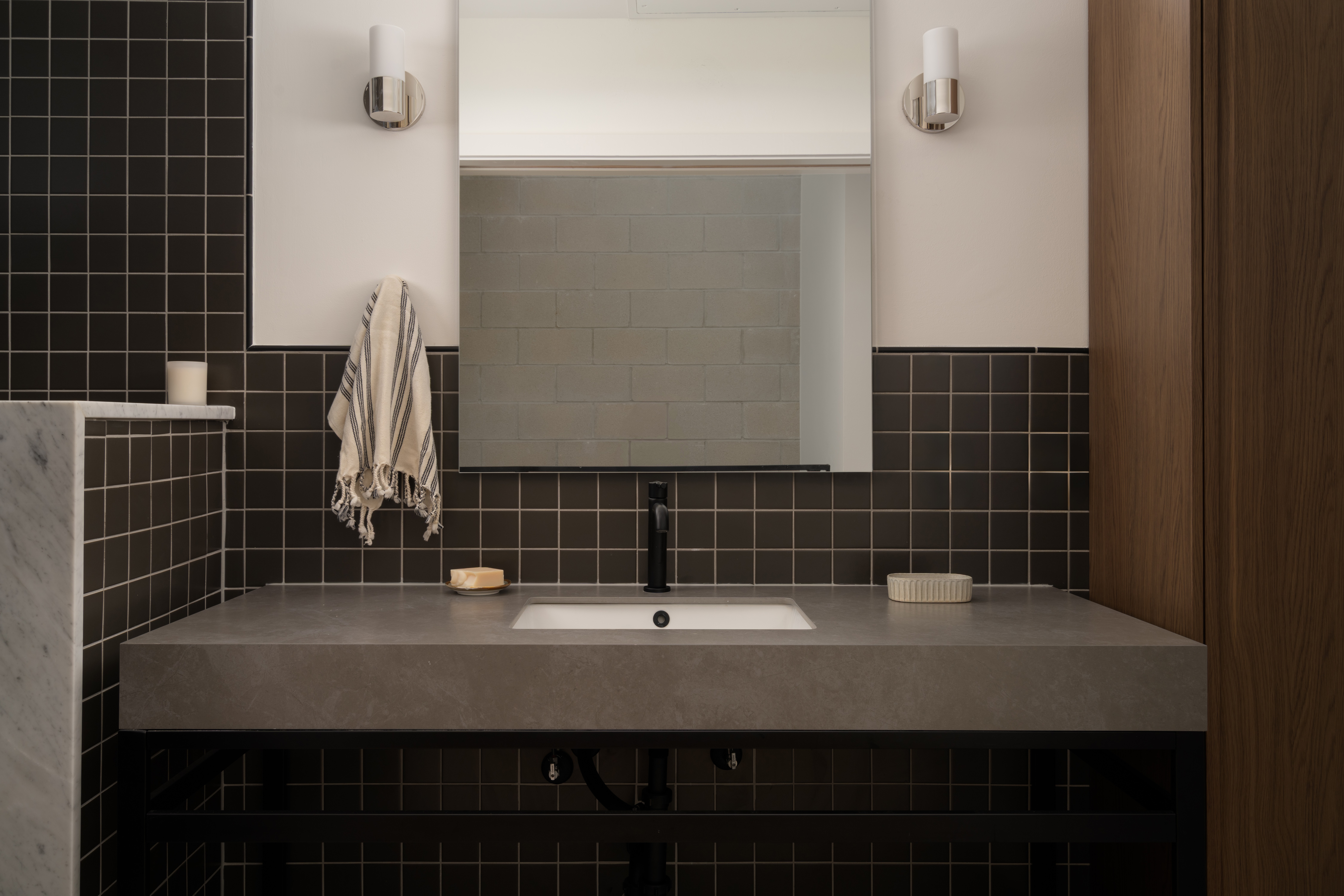 Bathroom vanity with a mirror, black tiled wall, and two wall-mounted lights, with a towel hanging on the left side.