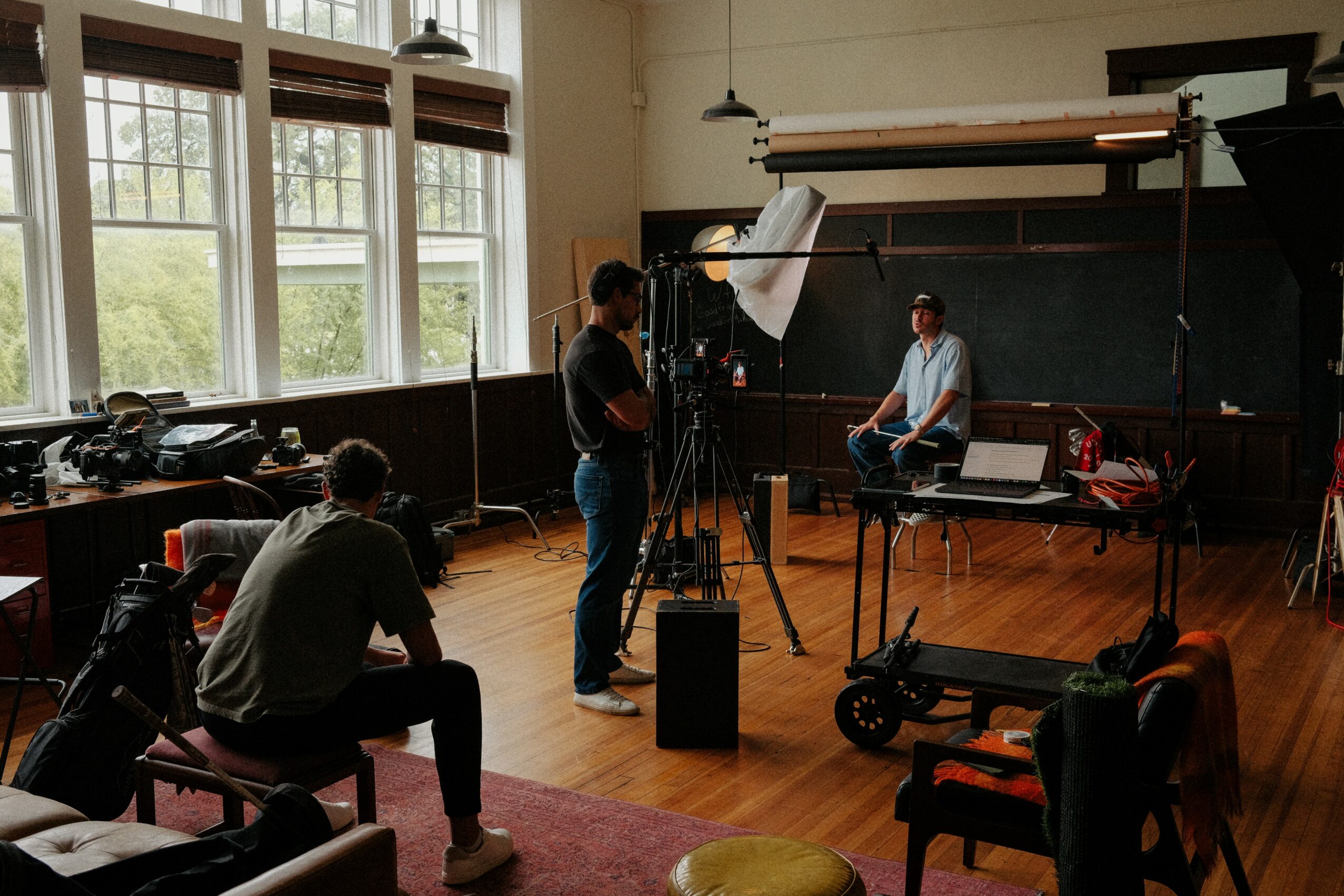 Person sitting on a chair in a room with large windows, filming a person sitting on a chair against a blackboard, with filming equipment around.