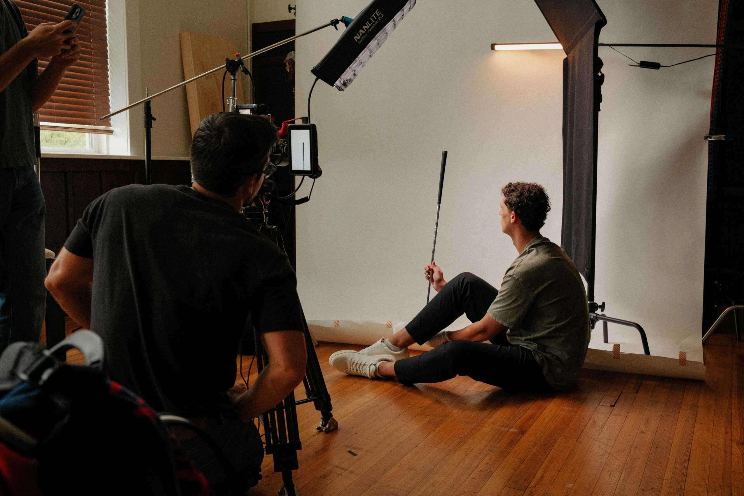 Person sitting on the floor holding a stick in a photography studio with lighting equipment and a white backdrop.