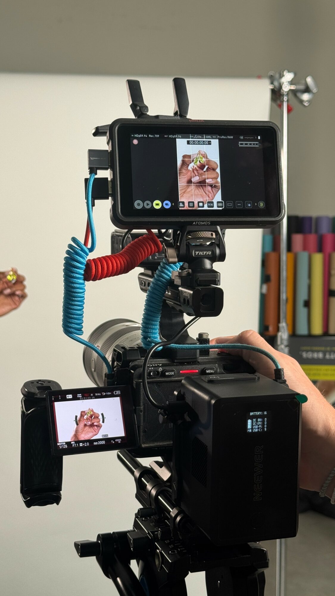 Camera setup capturing a person holding a small object, with a monitor displaying the close-up view, in a room with books.