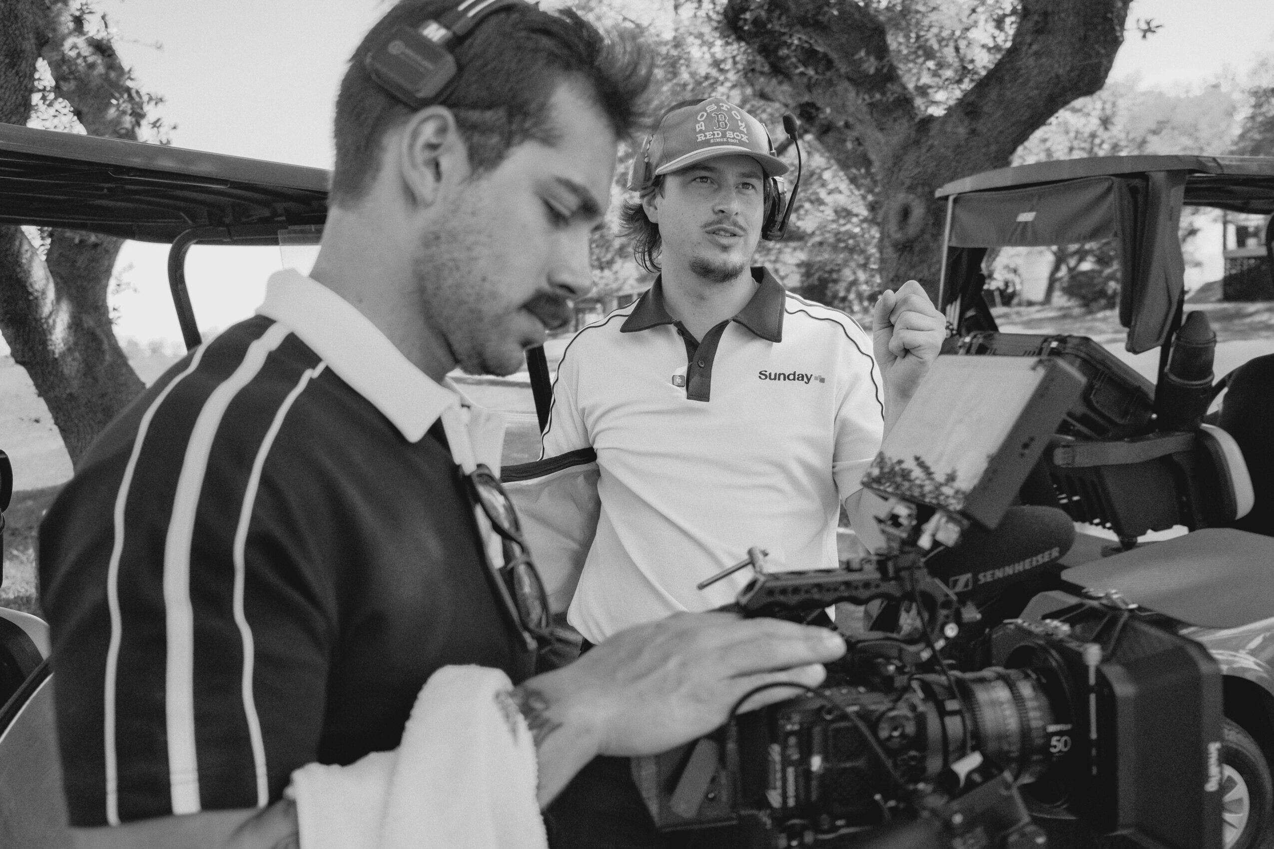 Two men outdoors, one operating a camera, the other observing, with trees in the background.
