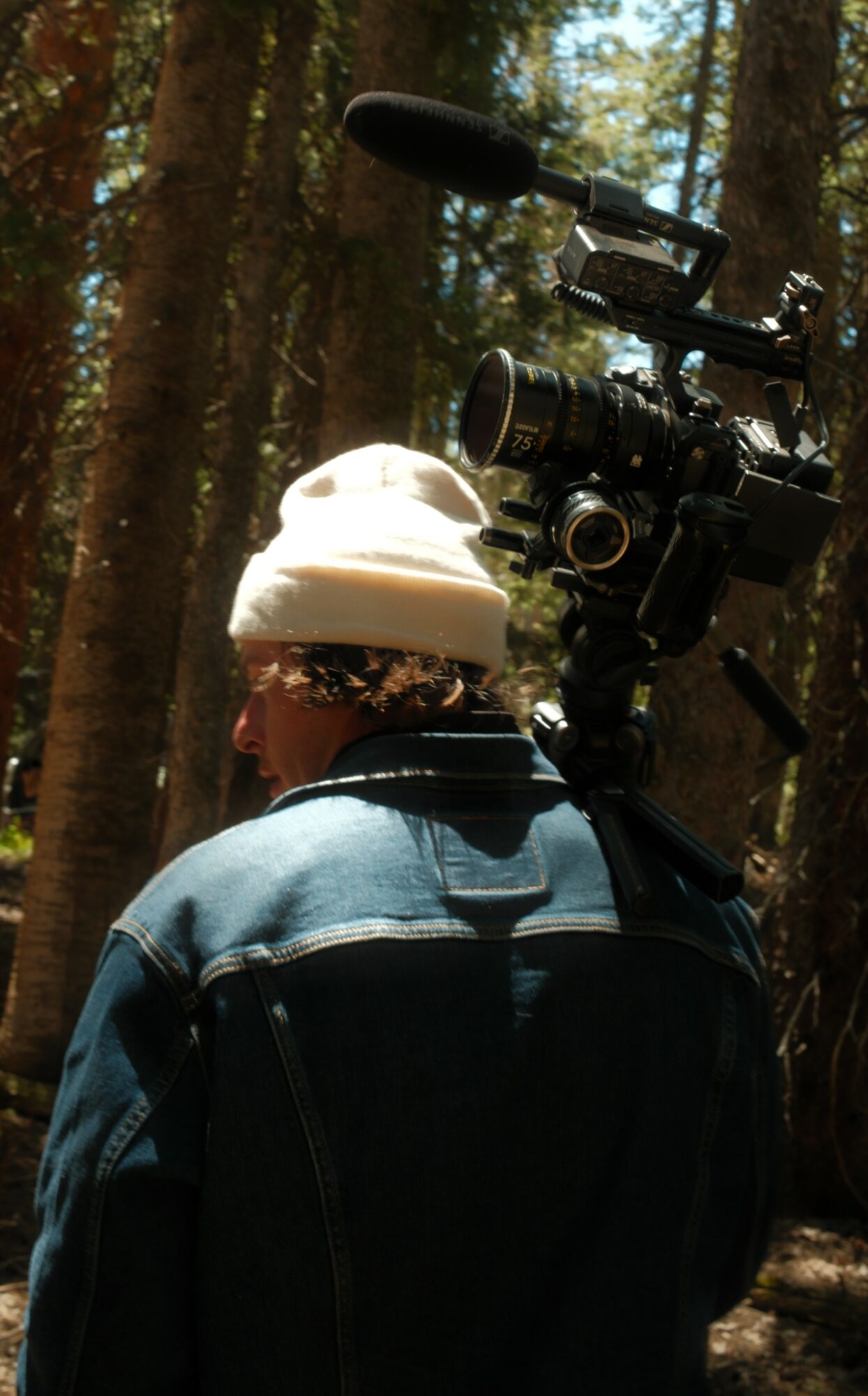 Person wearing a white hat and denim jacket in a forest with trees and sunlight.