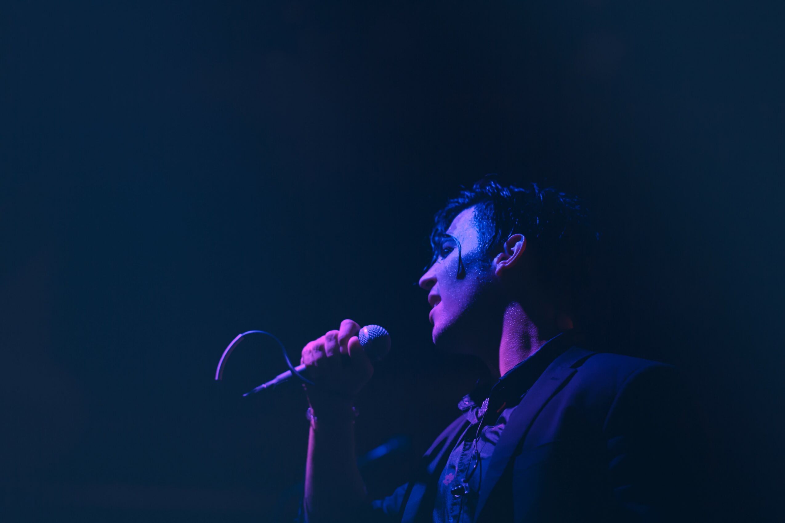 Person singing into a microphone on stage with dark background, illuminated by blue light.