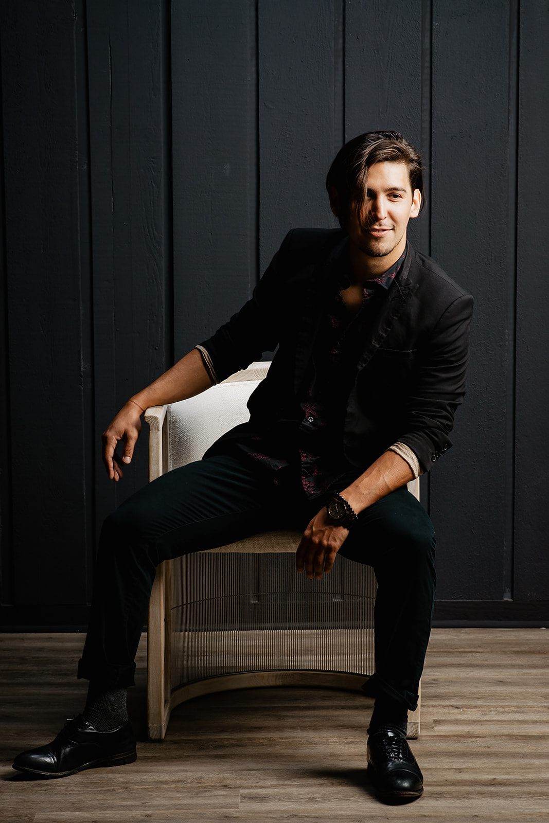 Man sitting on a chair against a dark wall, smiling, wearing a black suit and shoes, with a wooden floor.