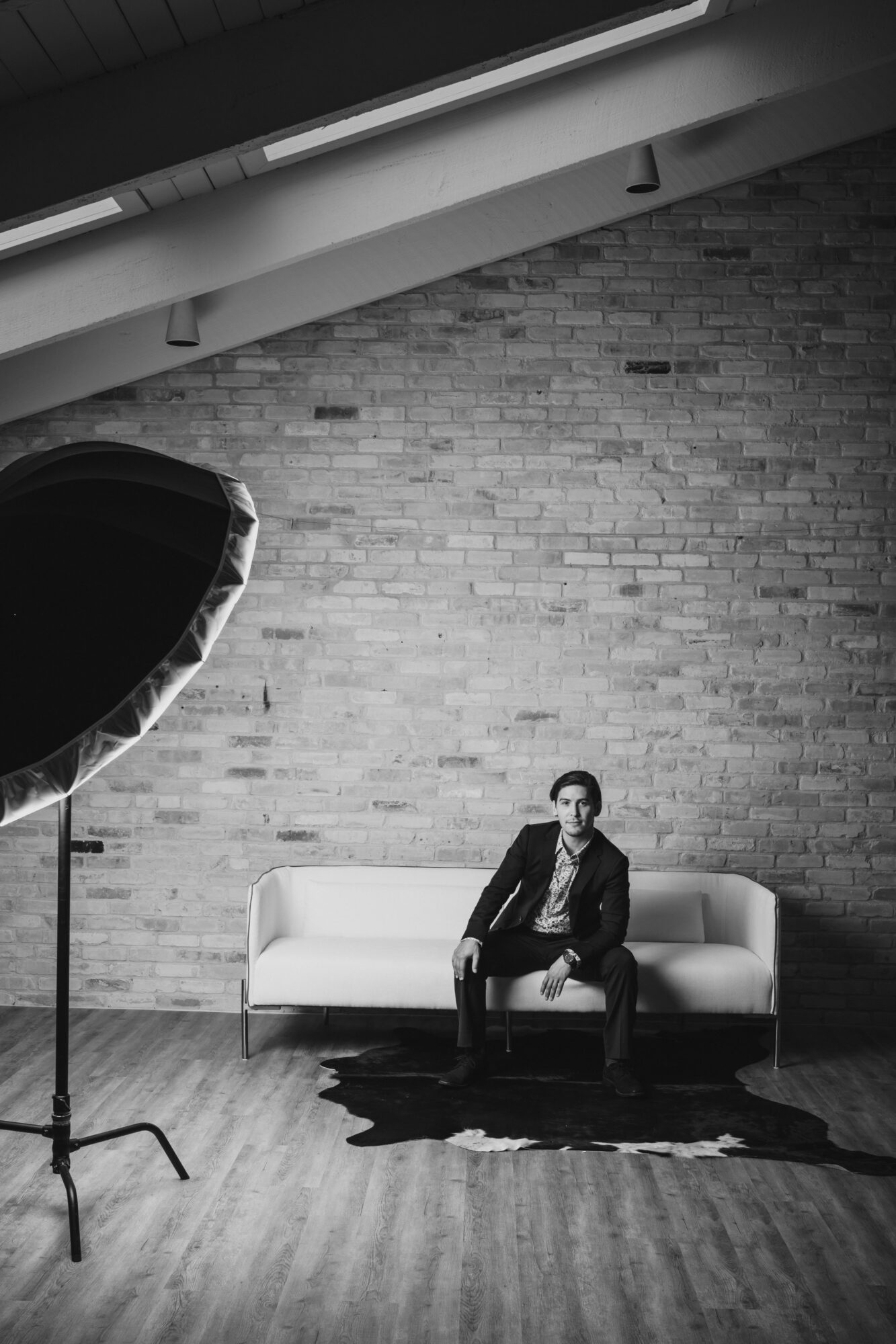 Person sitting on a white sofa in a room with a brick wall, wooden floor, and studio lighting, looking at the camera.