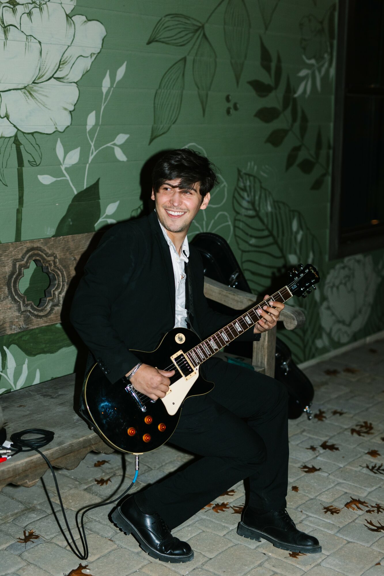 Young man with dark hair and glasses playing an electric guitar, sitting on a chair in front of a green floral mural.
