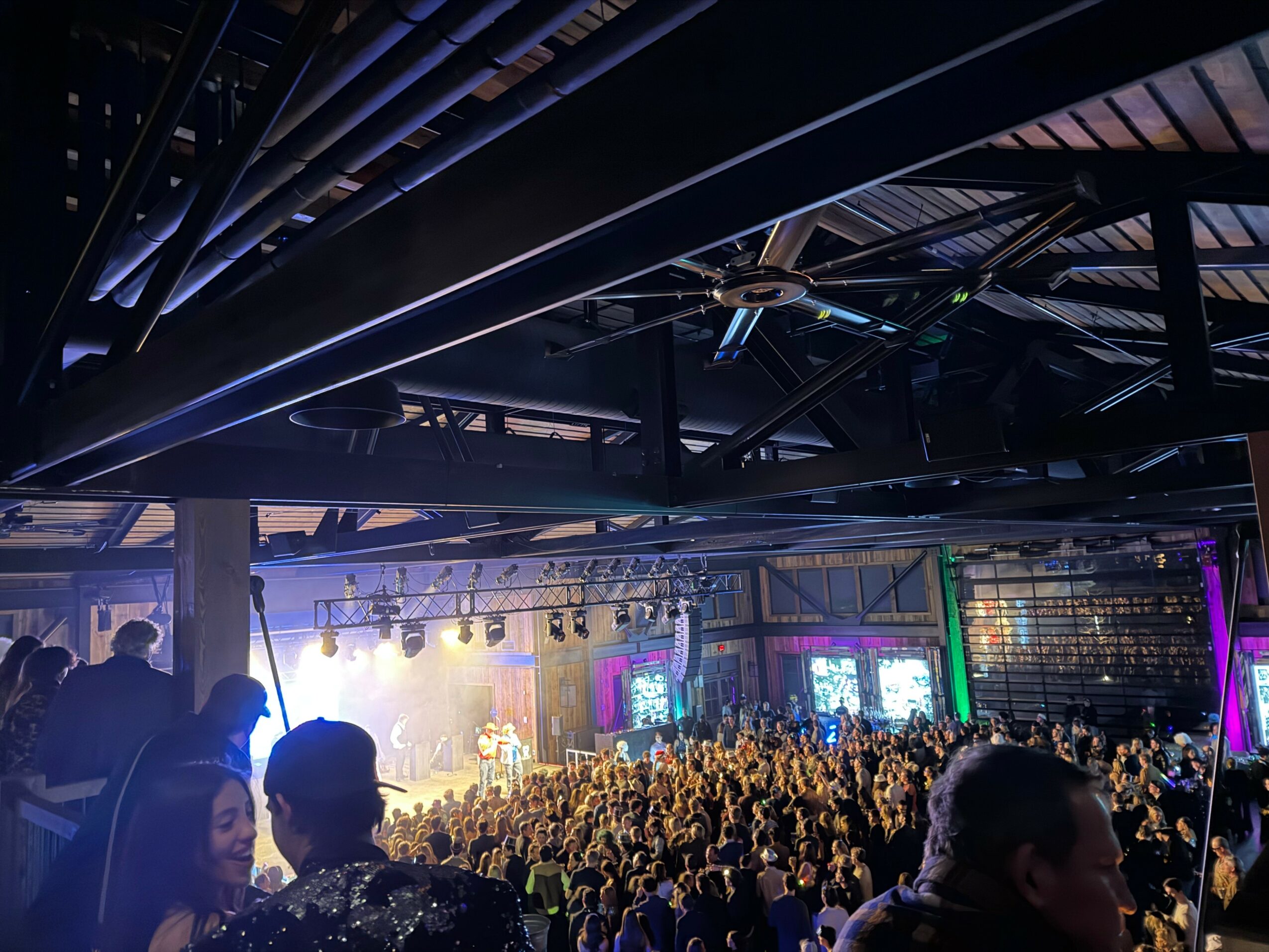 Crowd gathered in a large indoor concert venue with stage lighting and screens, viewed from above.