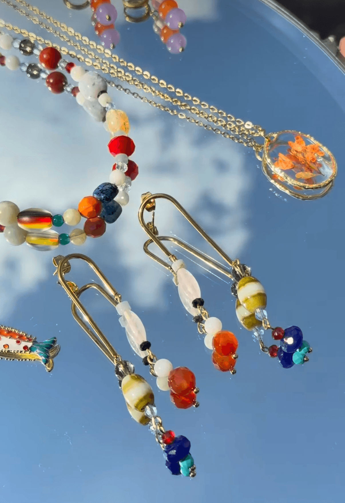 Three pairs of earrings with colorful beads and gold hooks, reflected on a mirror surface with a blue sky background.