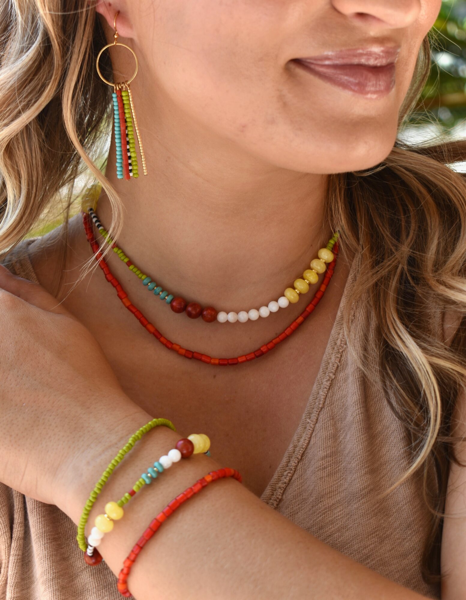 Close-up of a woman wearing colorful jewelry, including earrings, necklaces, and bracelets, with a beige top and wavy hair.