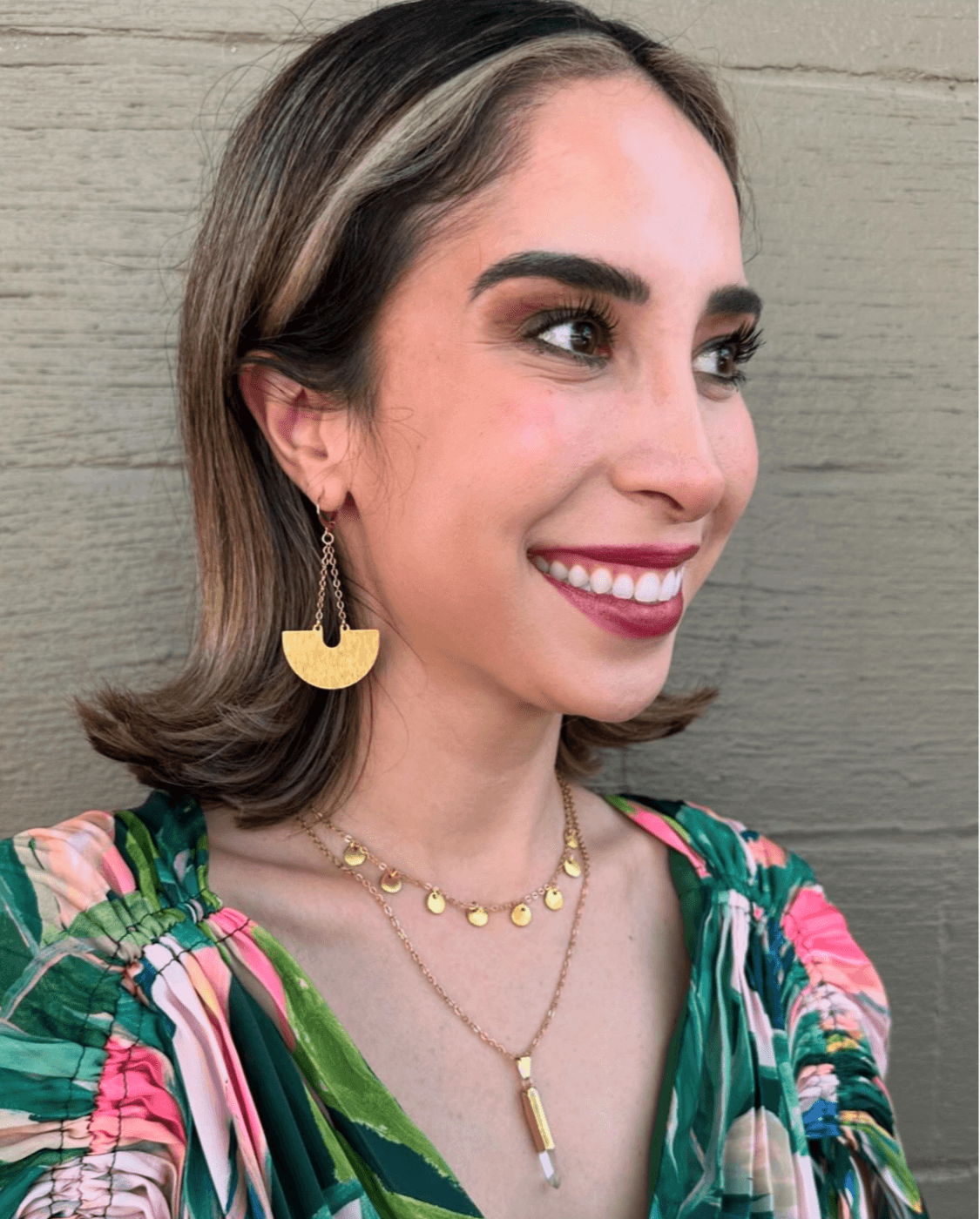 Woman with shoulder-length hair, smiling, wearing colorful floral top, gold earrings, and layered necklaces, standing against a wooden background.