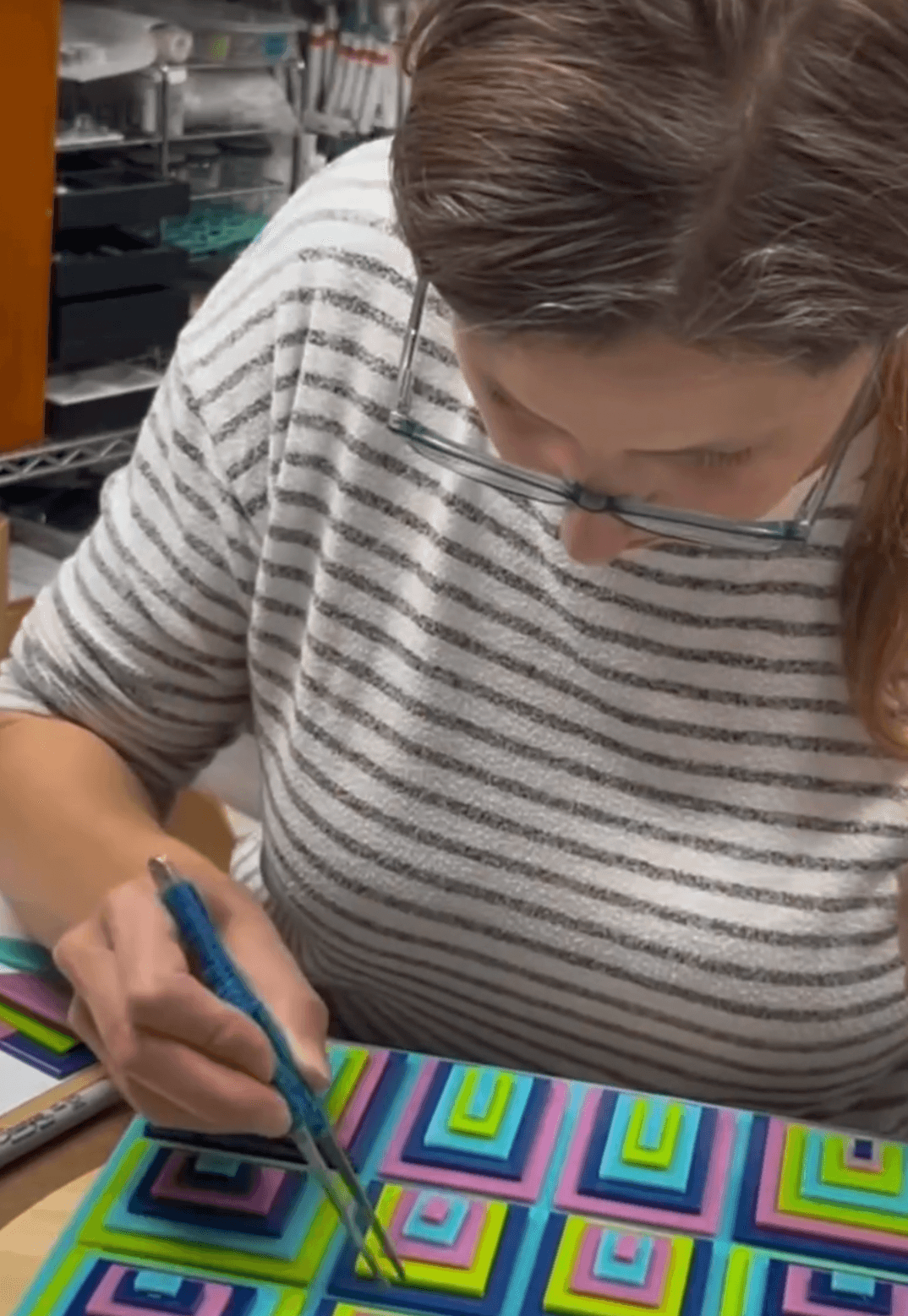 Person with glasses and striped shirt drawing on colorful geometric patterned paper, workspace with shelves in background.