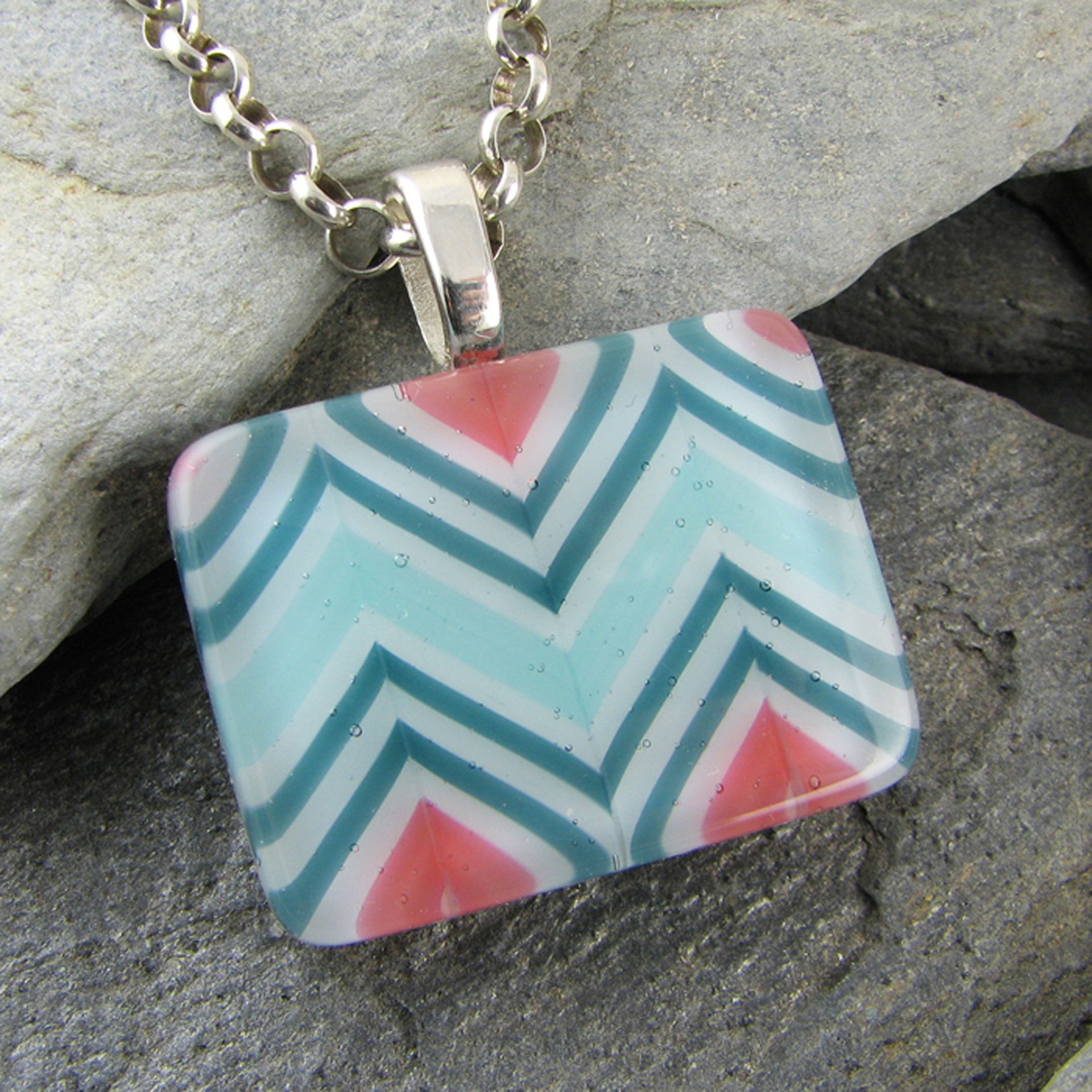 Rectangular pendant with a colorful zigzag pattern, attached to a chain, resting on rocks.