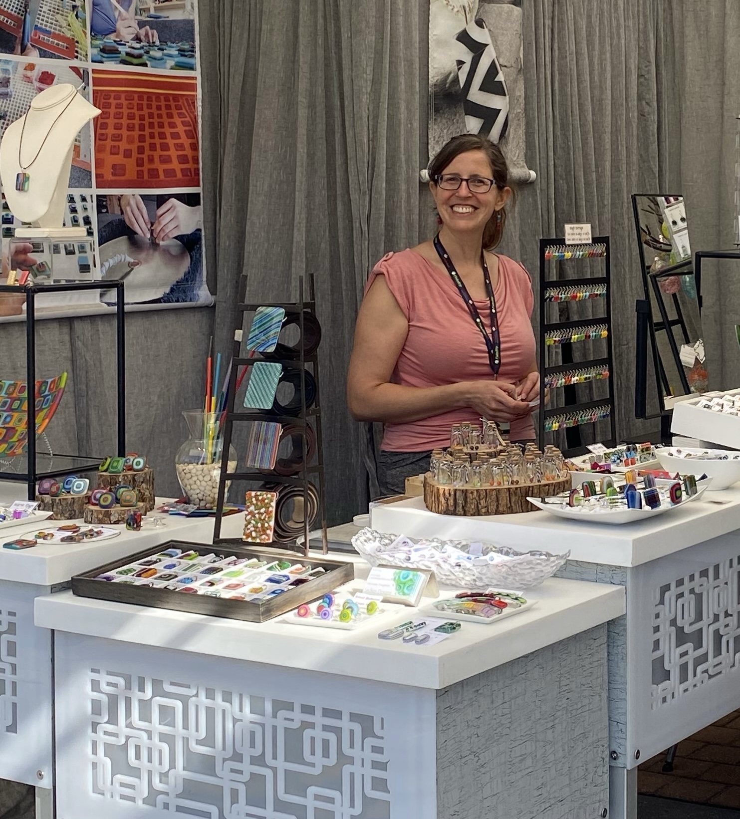 Woman standing behind a table with jewelry and craft supplies, smiling, in a booth with gray curtains and display racks.