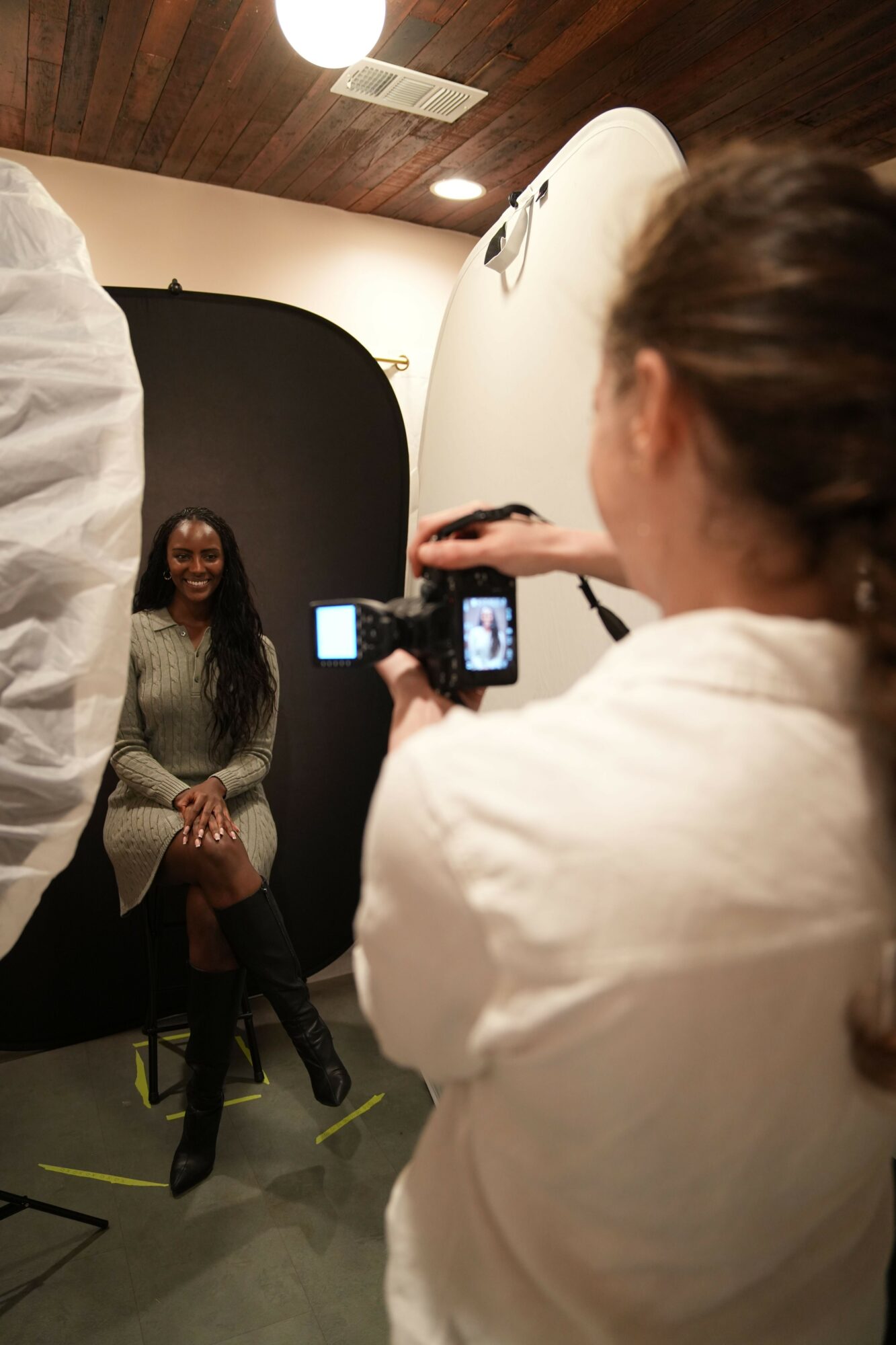 Person taking a photo of a woman standing against a black backdrop in a room with wooden walls and ceiling lights.