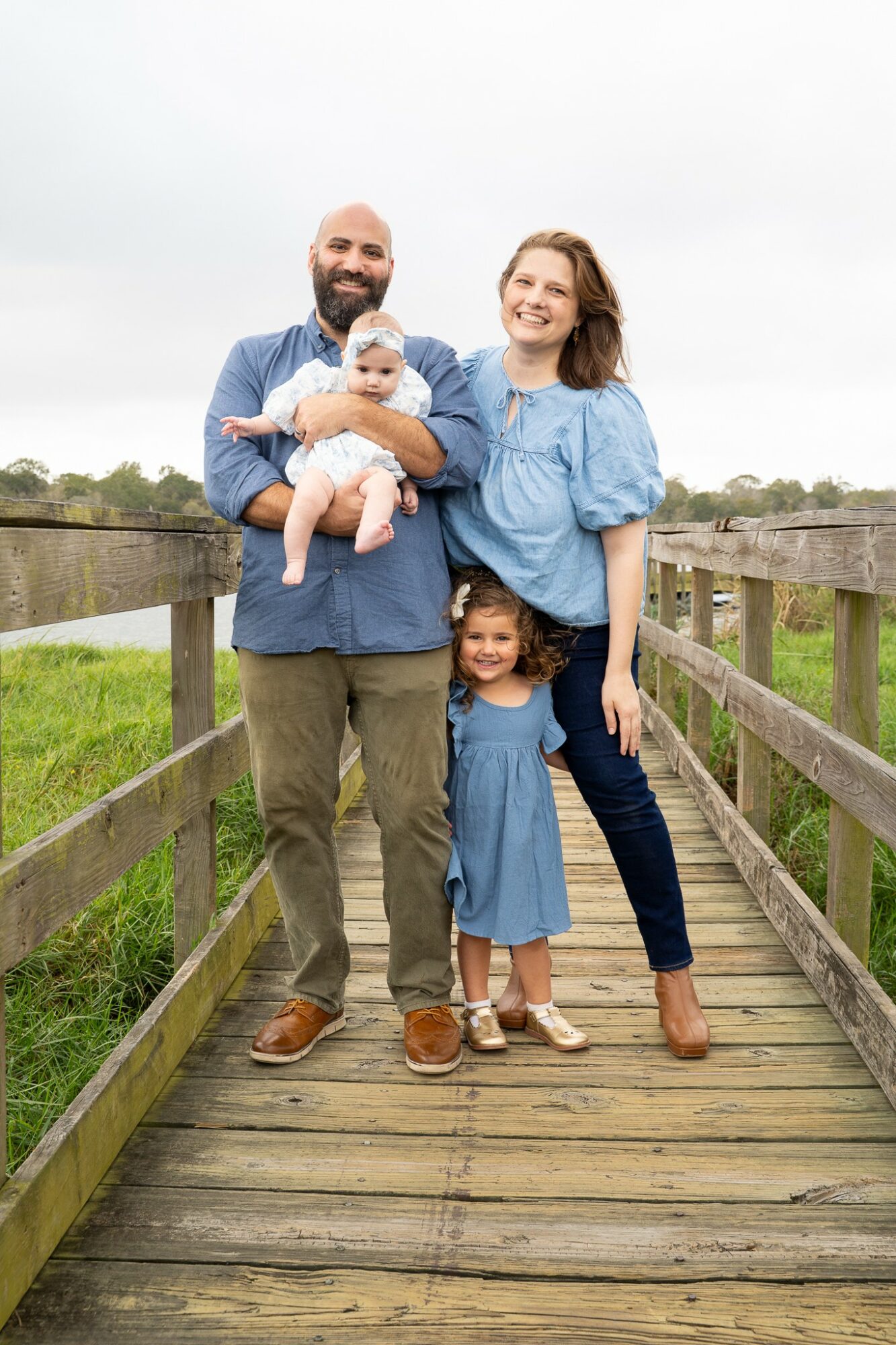 Family of four standing on a wooden bridge outdoors, smiling, with a cloudy sky and greenery in the background.