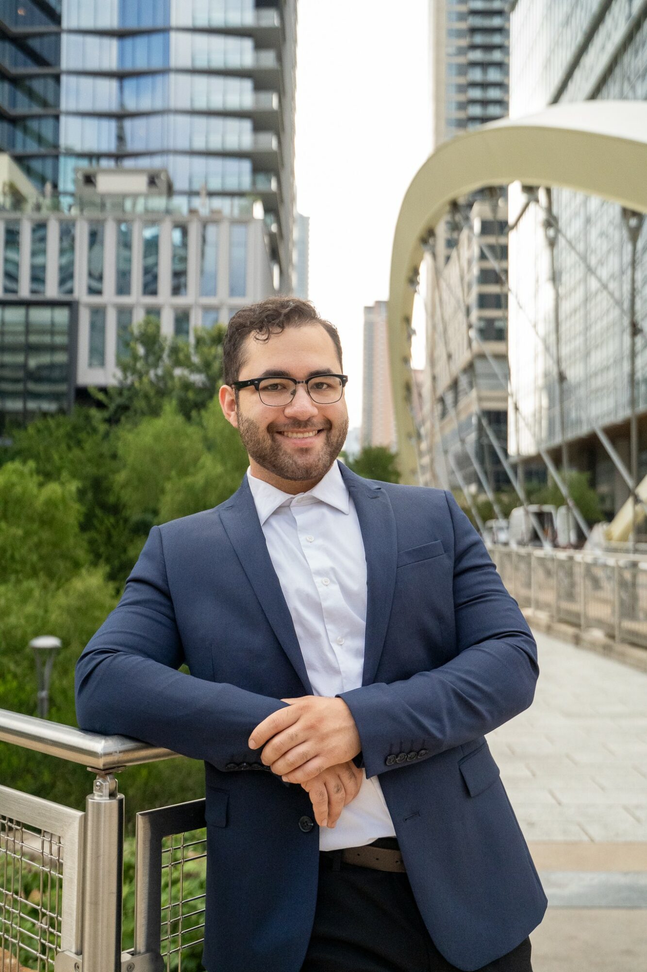 Man in a suit smiling outdoors in an urban setting with modern buildings and greenery.