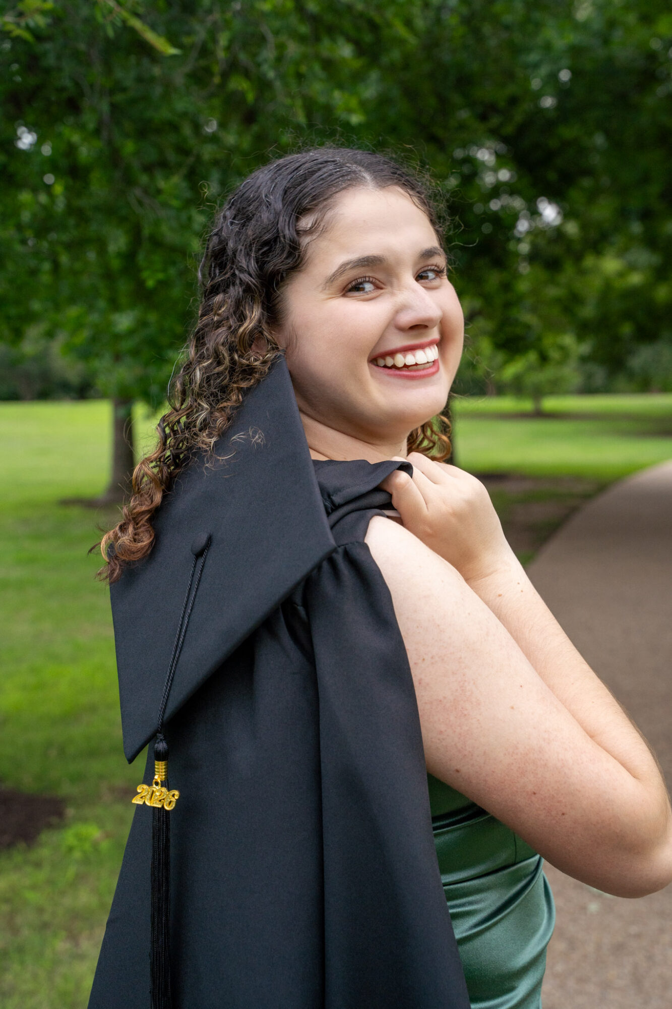 Young woman in graduation gown smiling outdoors, holding her gown over shoulder, trees and pathway in background.