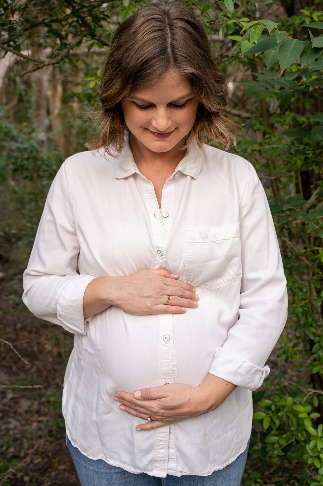 Pregnant woman with short brown hair in a white shirt standing outdoors among green foliage, smiling gently, hands on her belly.