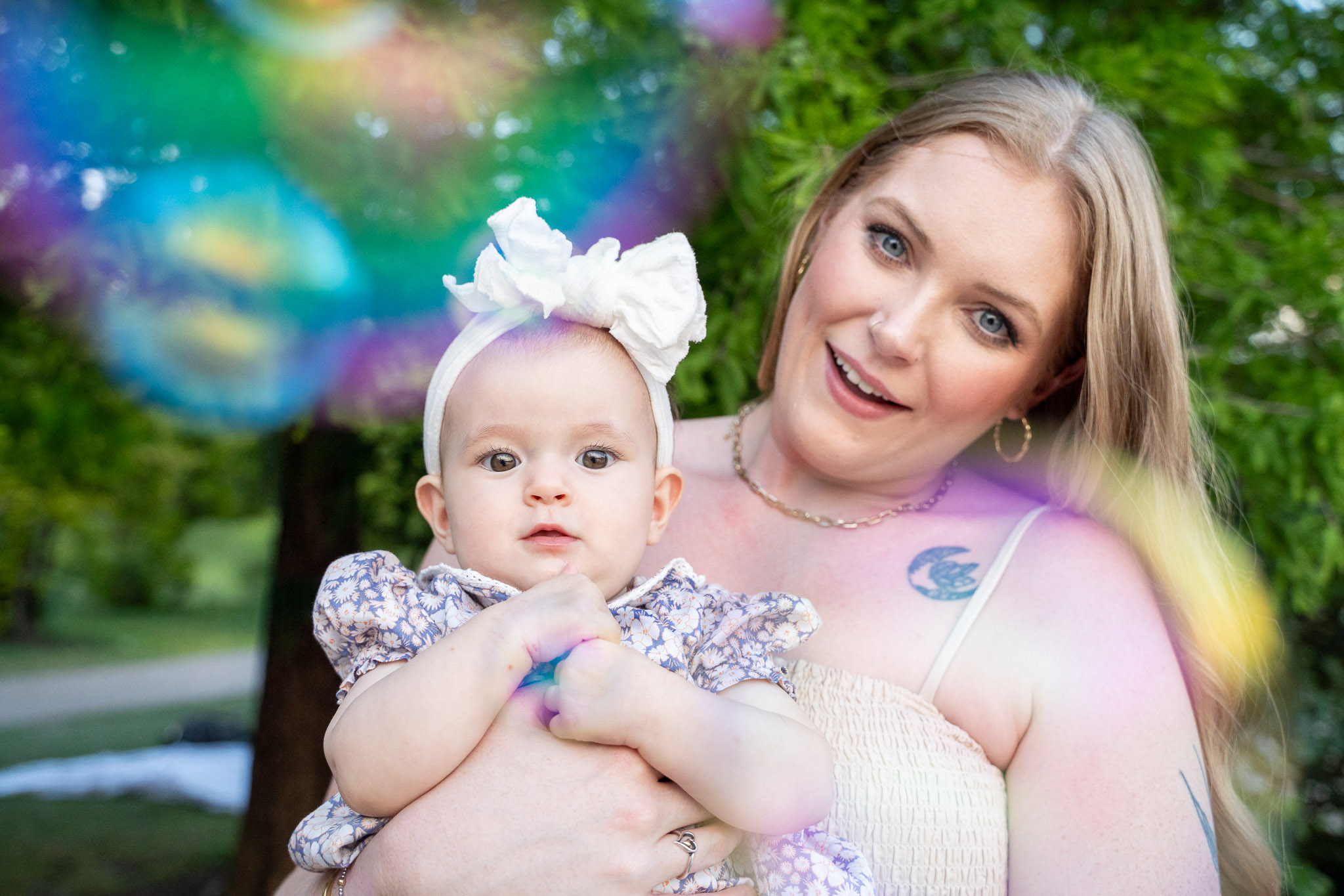 Woman holding a baby outdoors with colorful light effects, trees, and greenery in the background.
