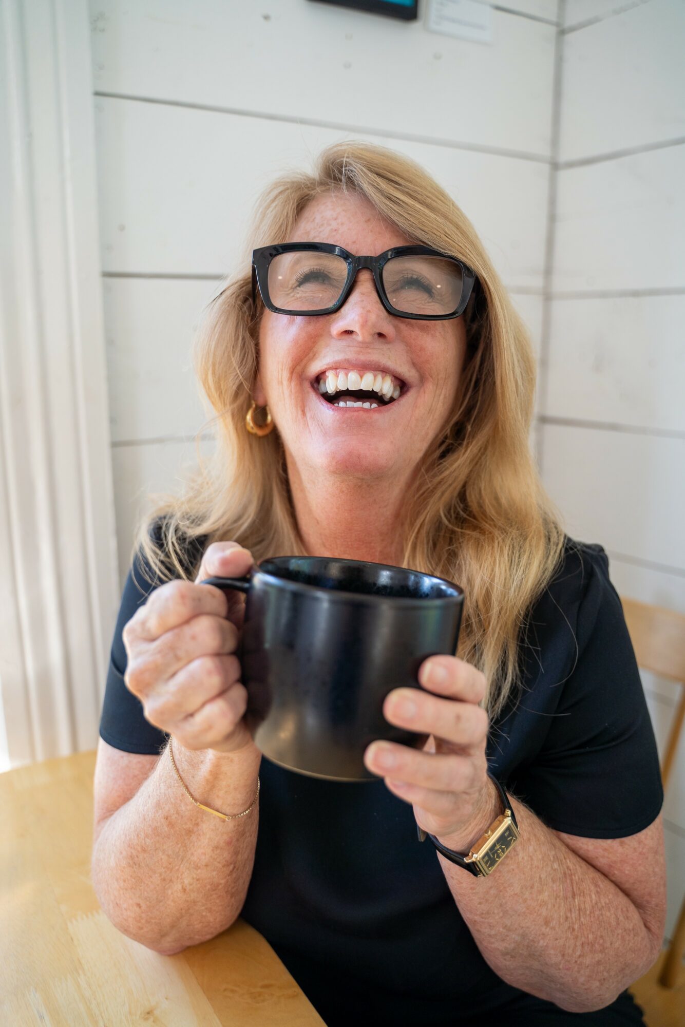 Woman with glasses smiling and holding a black mug, sitting at a table indoors.