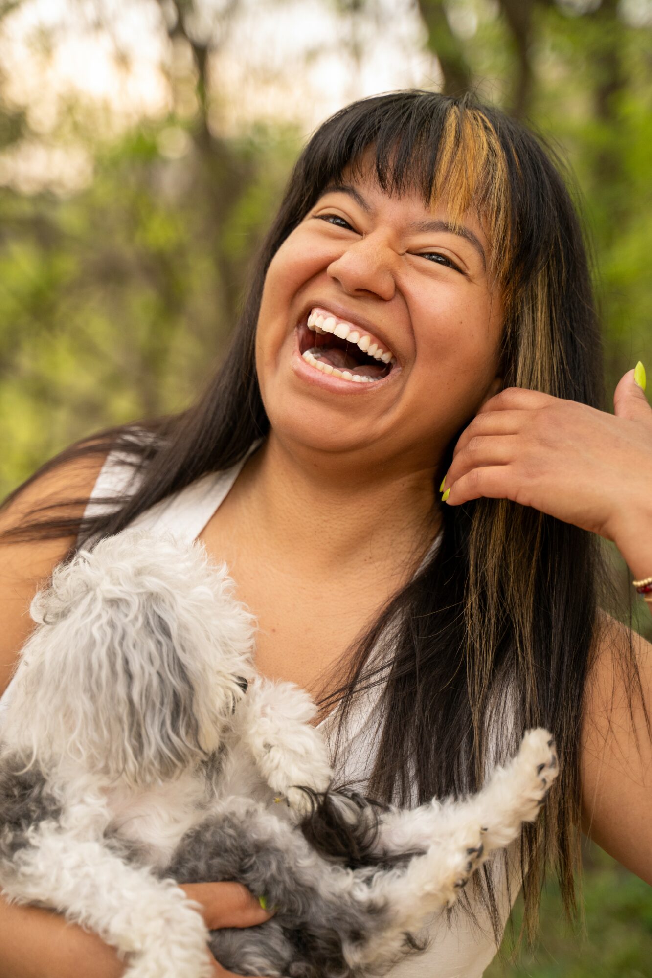 Woman laughing outdoors holding a small dog, trees in background, smiling with eyes closed.