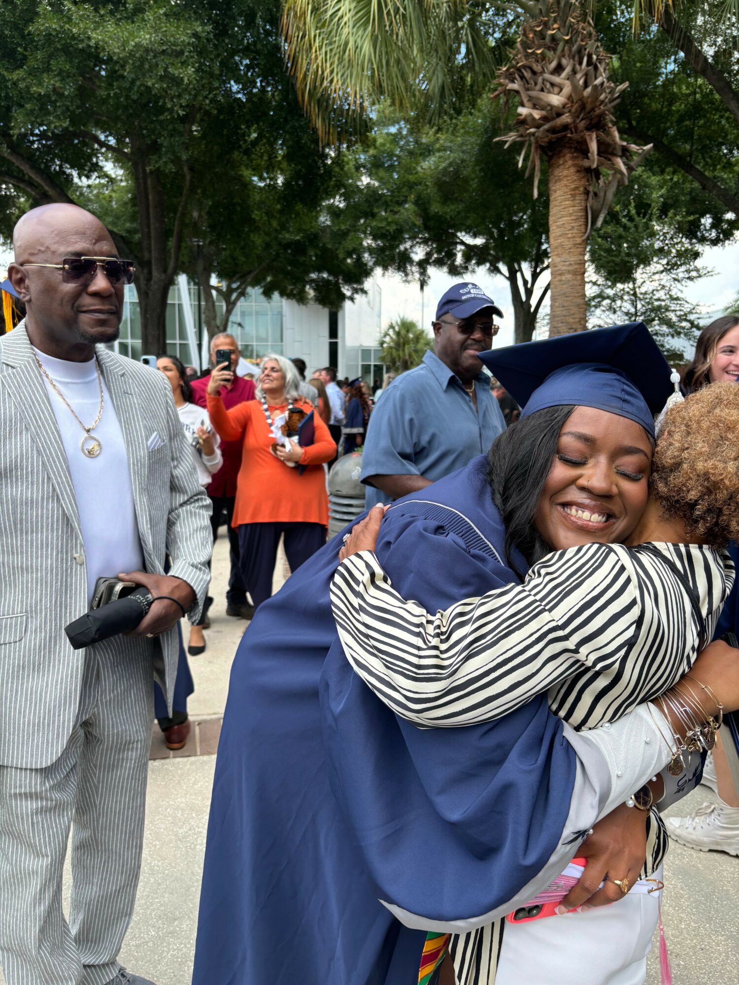 Person in graduation gown hugging another person outdoors, smiling, with a group of people and trees in background.