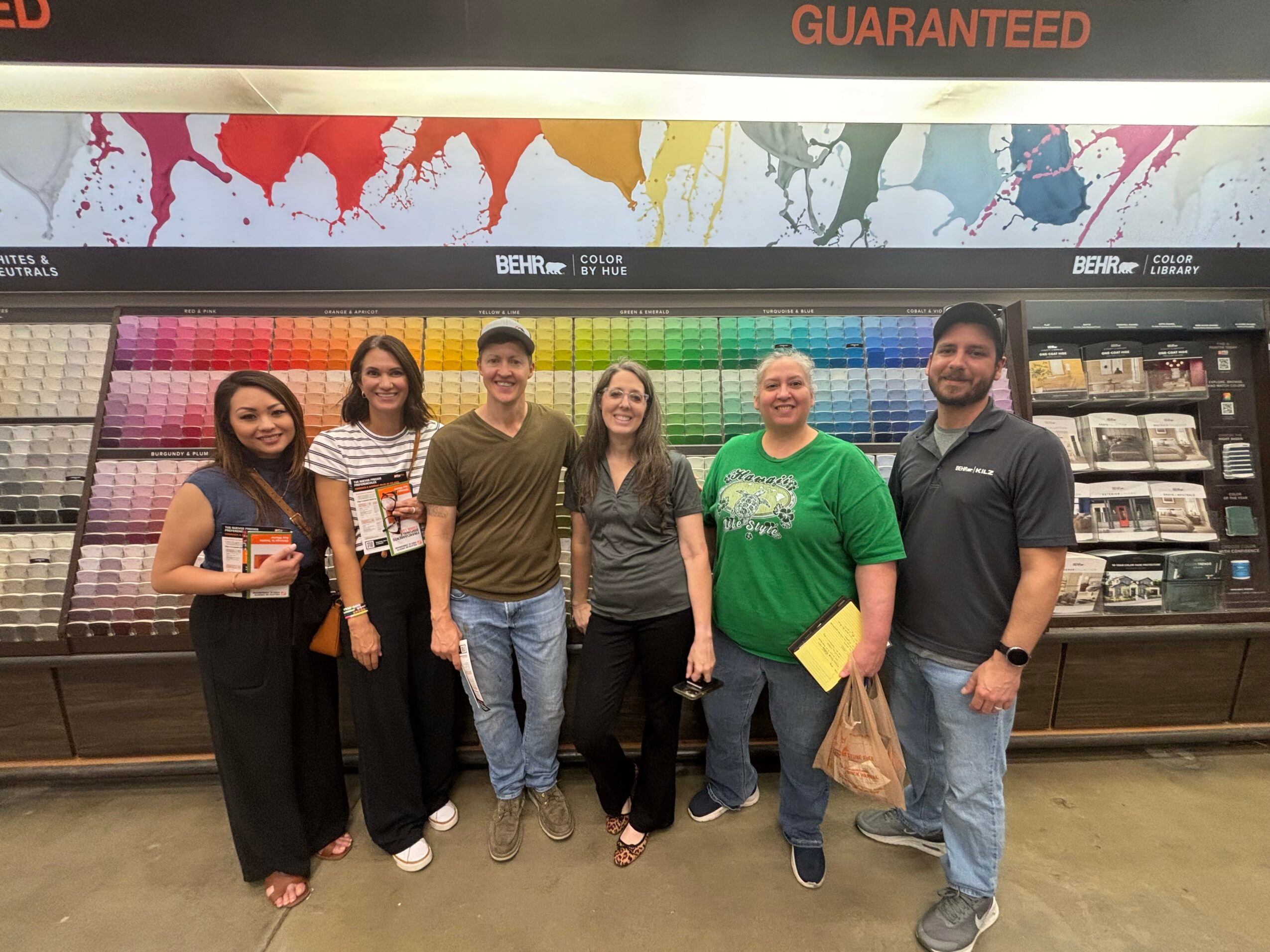 Group of six people standing in front of a colorful paint display in a store, smiling at the camera.