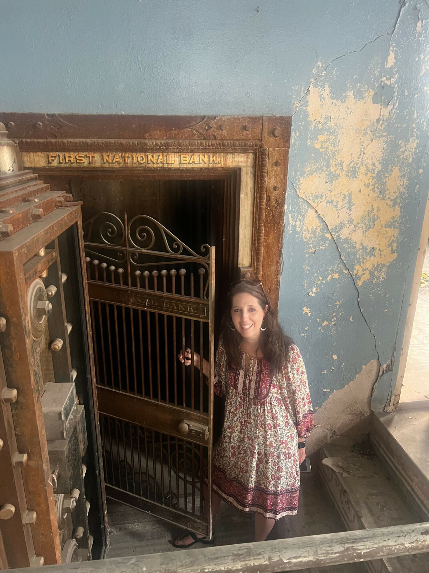 Woman standing near a decorative metal gate in a worn building with peeling paint and a wooden door frame.