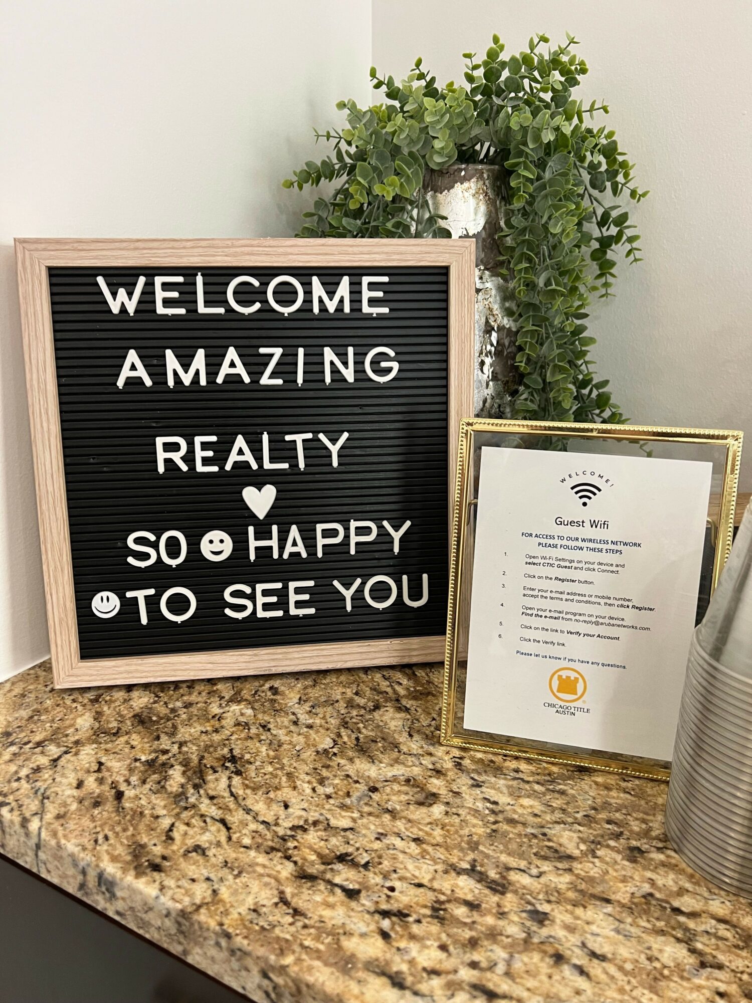 Letter board with welcoming message, plant, and framed certificate on a granite countertop.