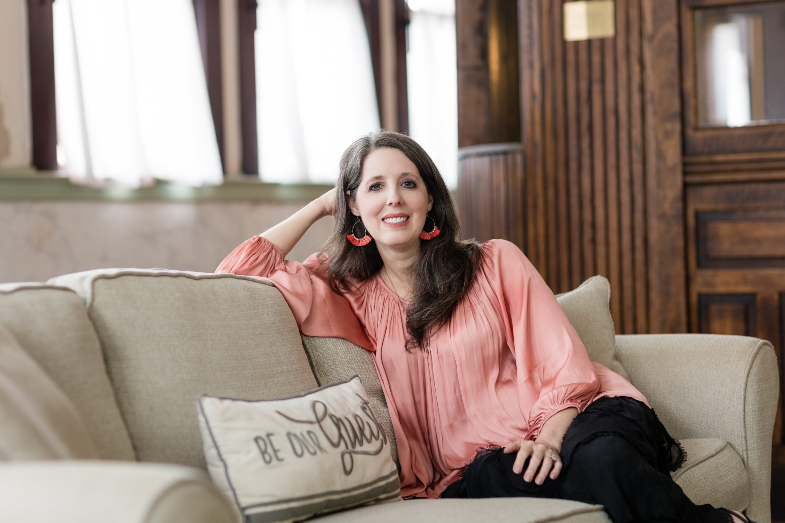 Woman with long dark hair wearing a pink blouse sitting on a beige sofa in a cozy room with wooden walls and windows.