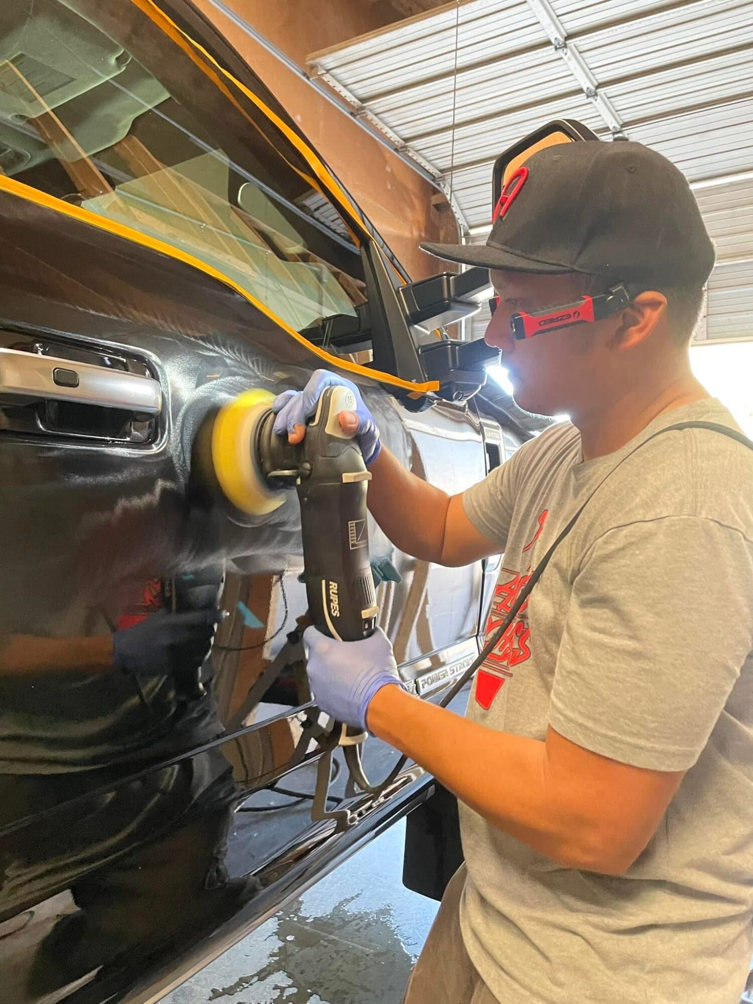 Person polishing a black car with a power buffer, wearing safety glasses, gloves, and a cap in a garage.