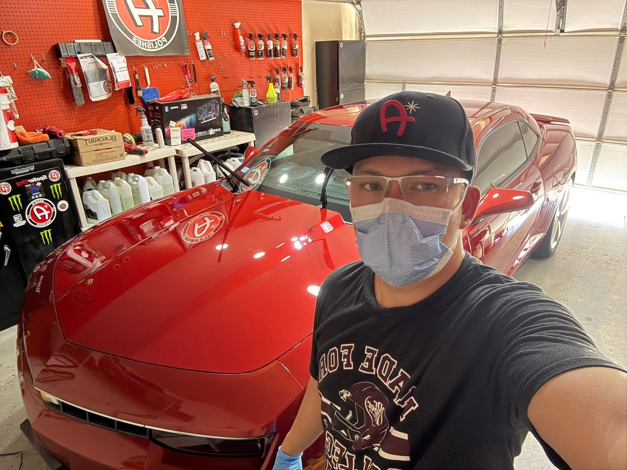 Person wearing glasses, a face mask, and a black cap taking a selfie in a garage with a red sports car behind them.