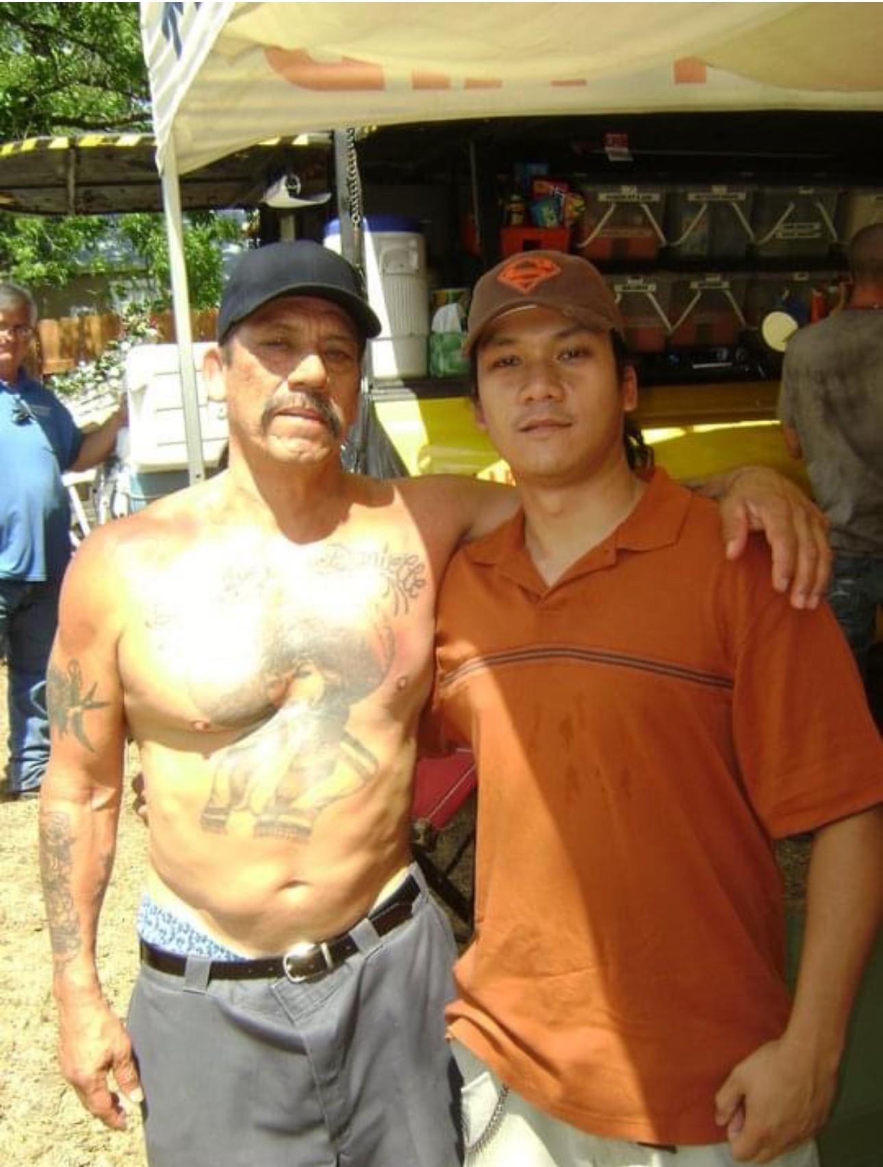 Two men standing outdoors, one shirtless with tattoos and the other in a brown shirt and cap, under a canopy.