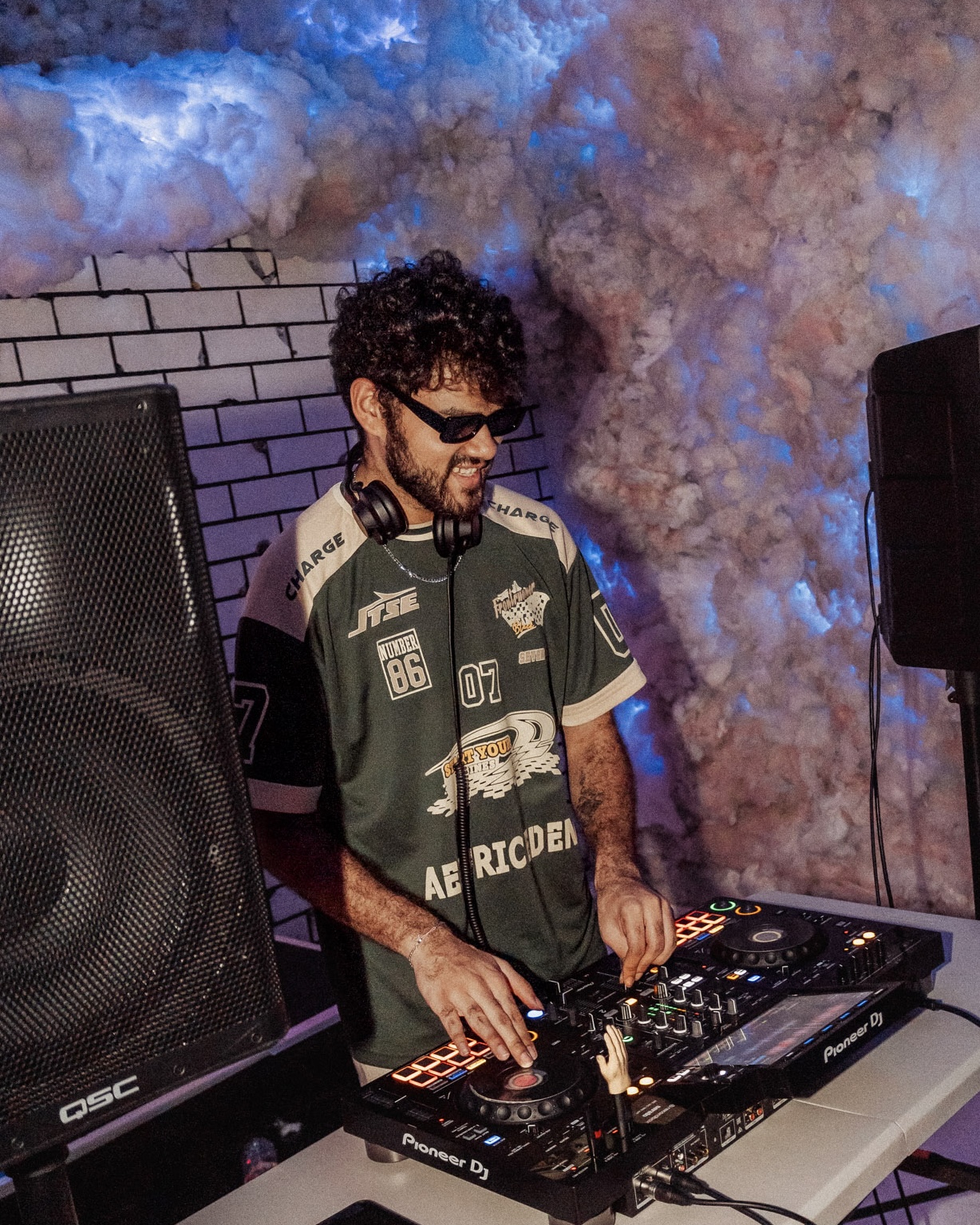 DJ with curly hair and sunglasses operates a DJ controller, surrounded by speakers and smoke effects, in a club setting.