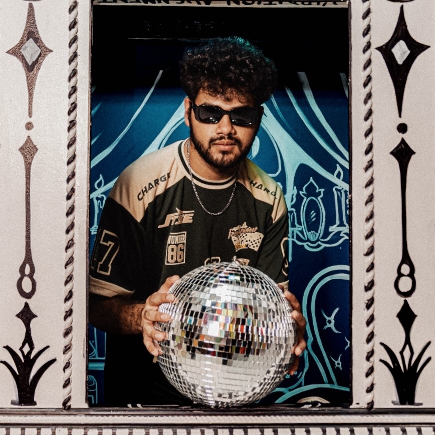 Young man with sunglasses and curly hair holding a disco ball, standing in front of a decorative background.