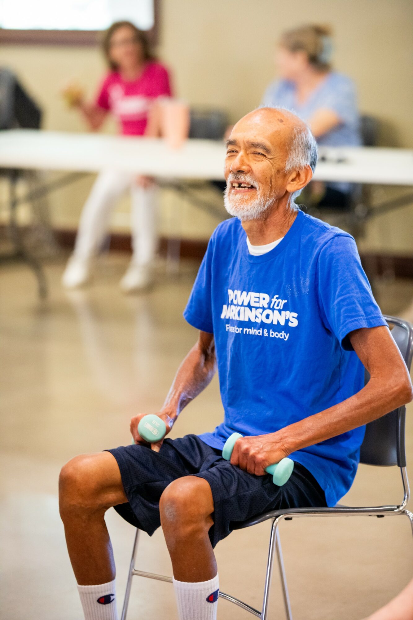 Elderly man sitting on a chair, smiling, wearing a blue T-shirt and black shorts, holding a small object in his hand.