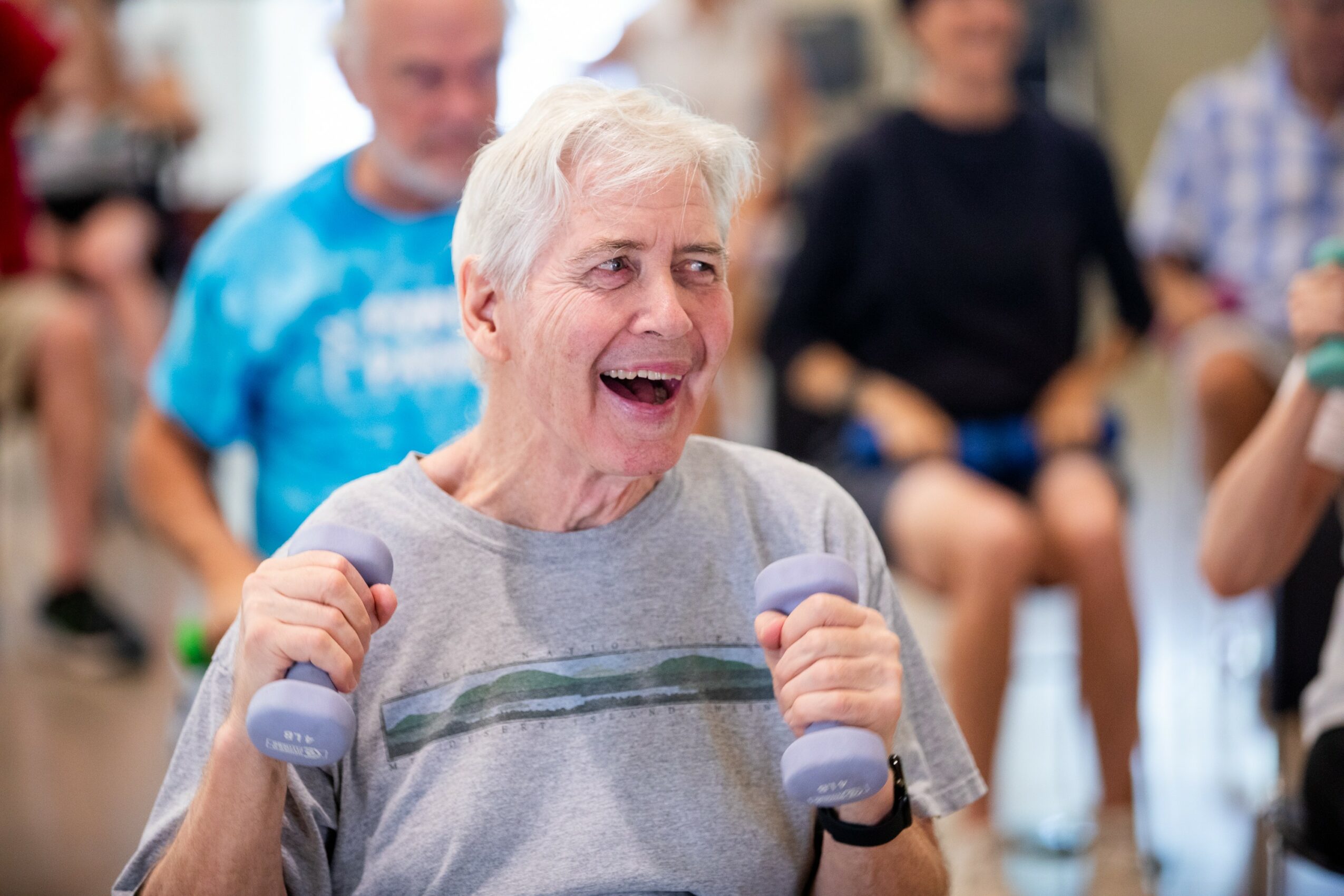 Older woman smiling and exercising with small dumbbells in a group class, surrounded by people, indoors.