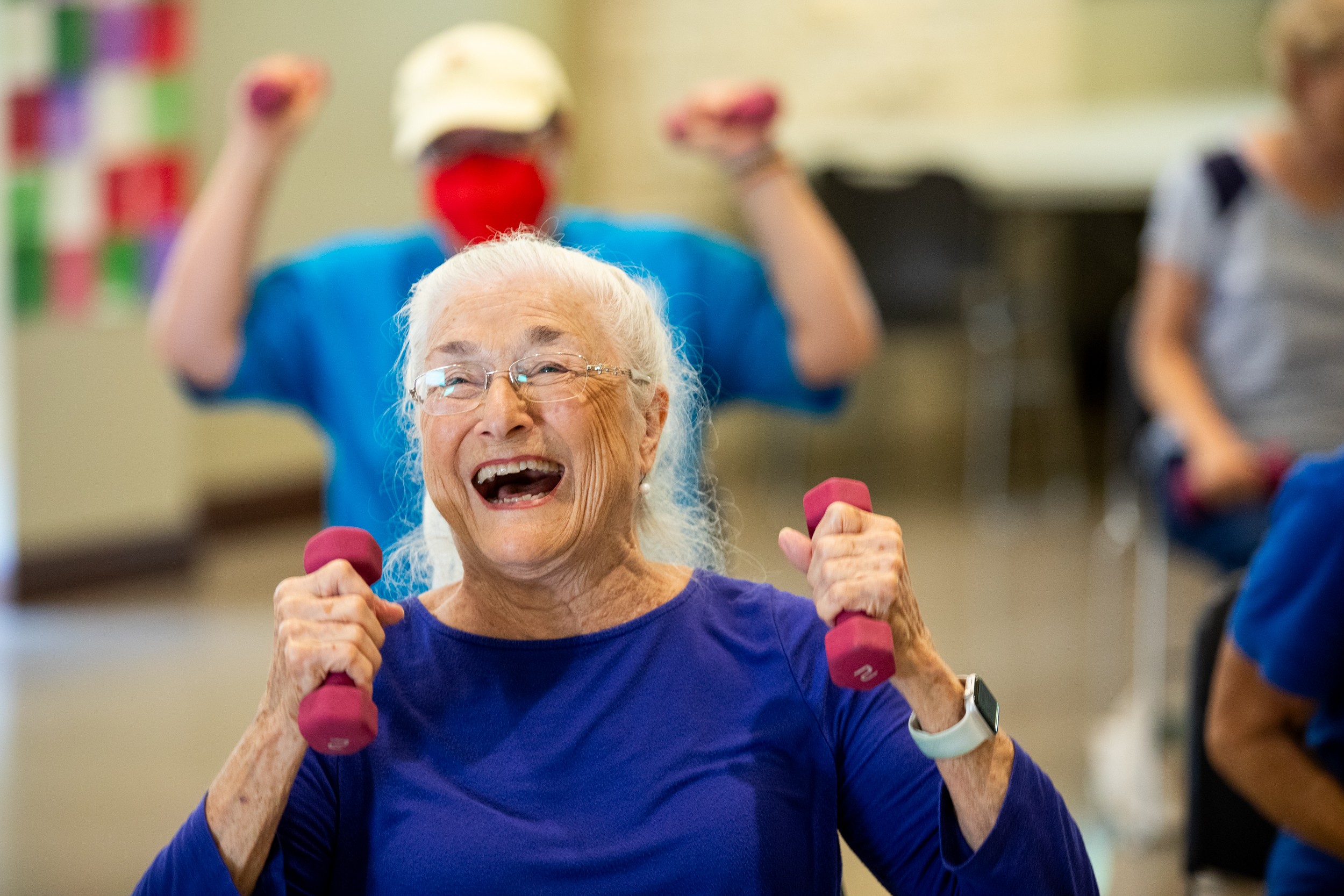 Elderly woman smiling and exercising with small pink dumbbells in a group activity, others in the background.