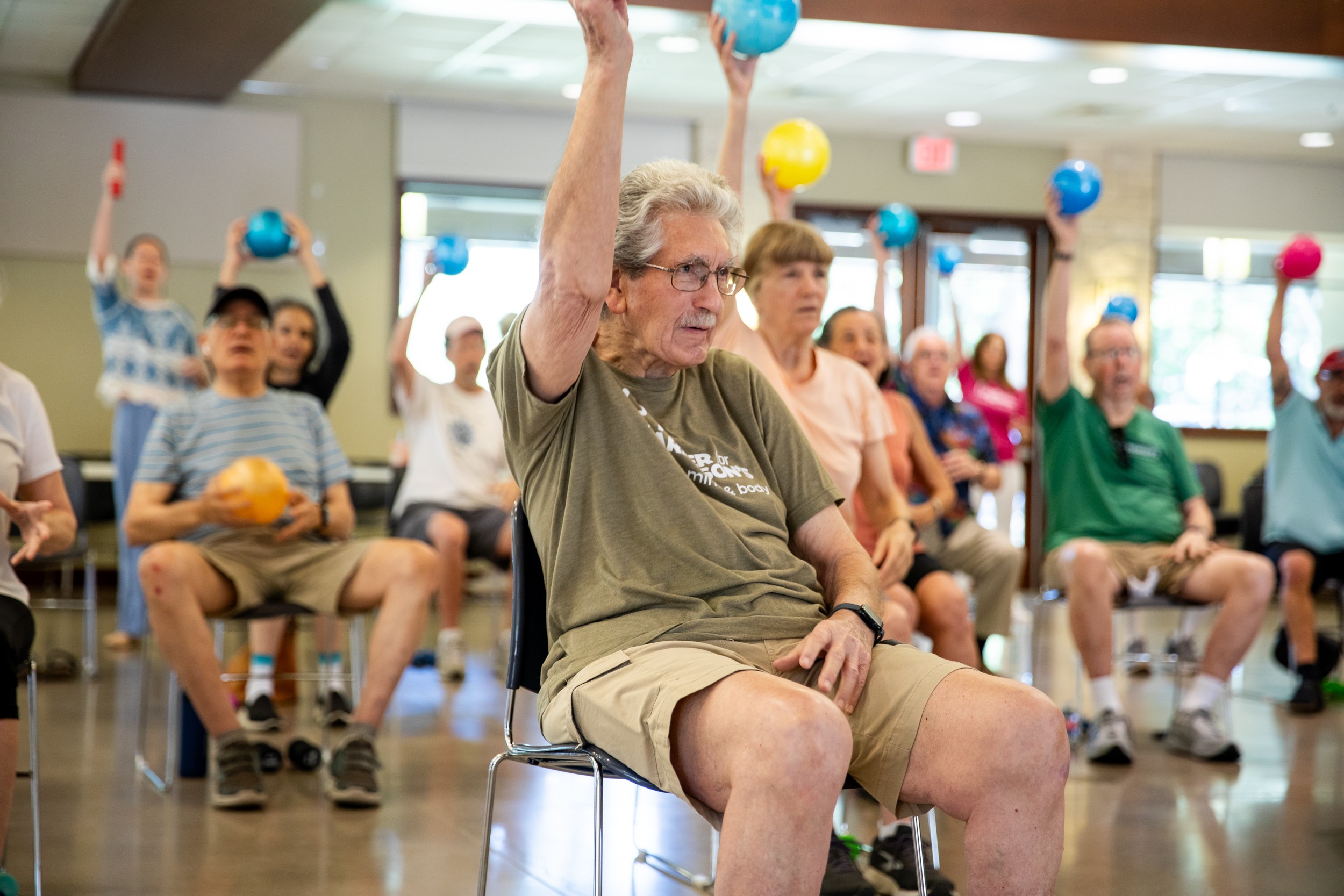 Older man raising a blue ball during a group exercise class with seated participants holding colorful balls, in a bright room.