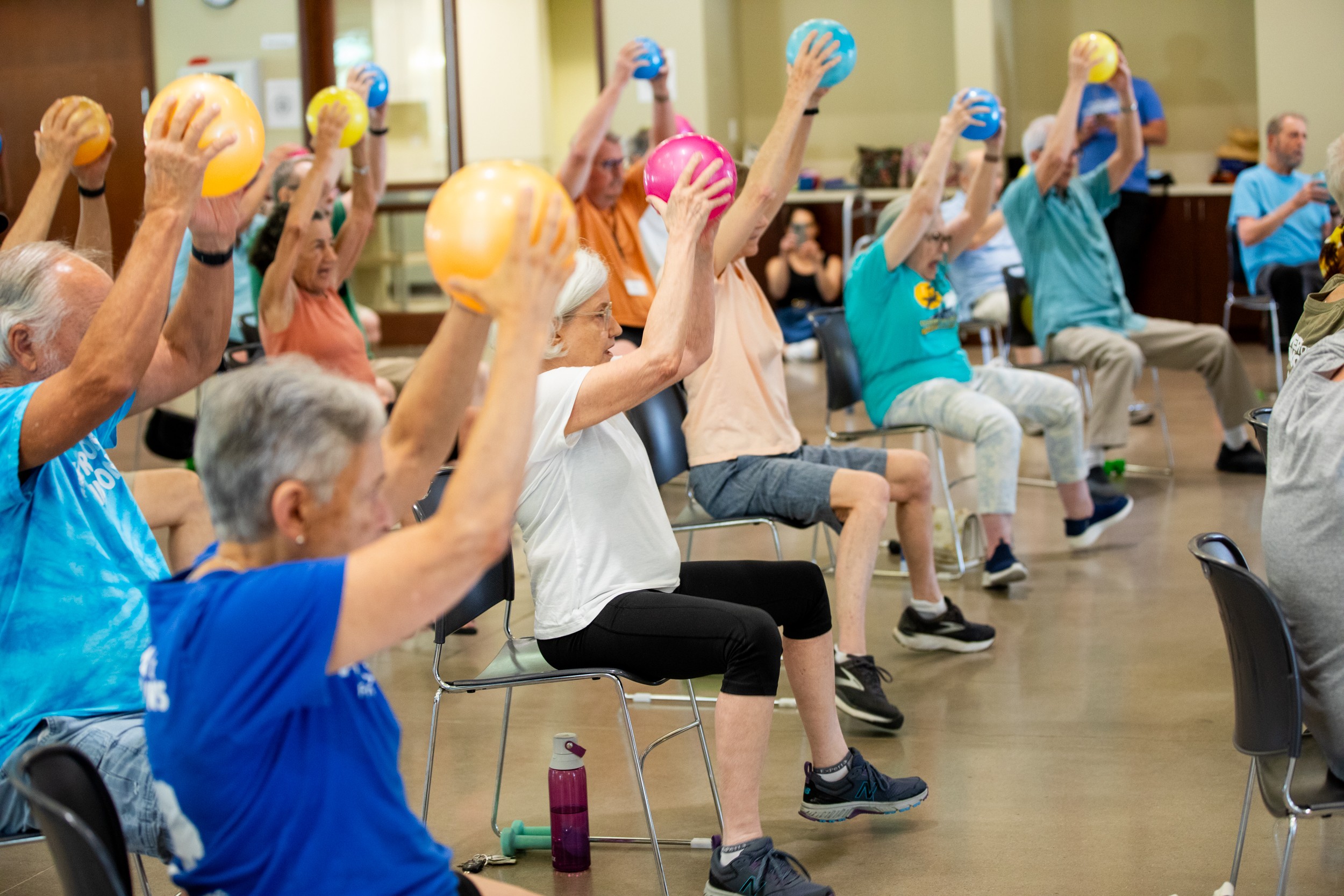 Group of elderly people participating in seated exercise class, holding small balls overhead, in a spacious room.