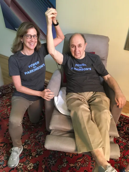 A woman and a man, both wearing Power Parkinson's shirts, smiling; the man is seated in a chair, raising a fist.