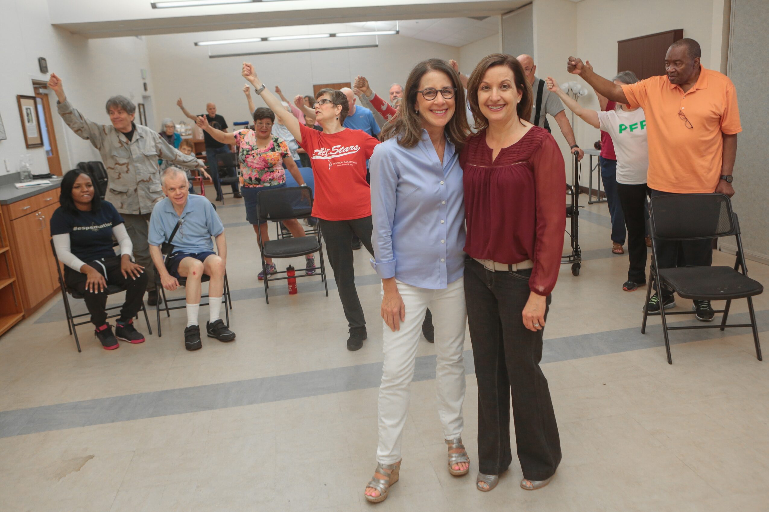 Group of diverse people in a room, some seated and some standing, with two women smiling in the foreground.