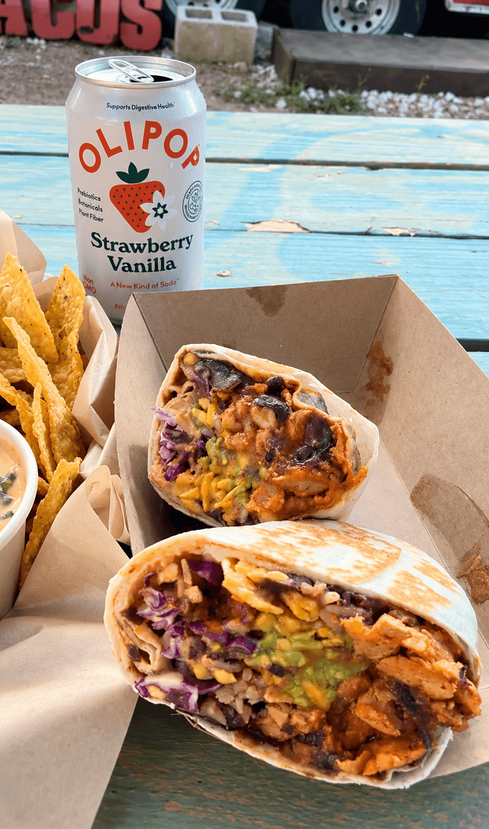 Half-eaten burrito with colorful filling on a tray, can of Olipop Strawberry Vanilla soda, and crispy chips on a blue table.