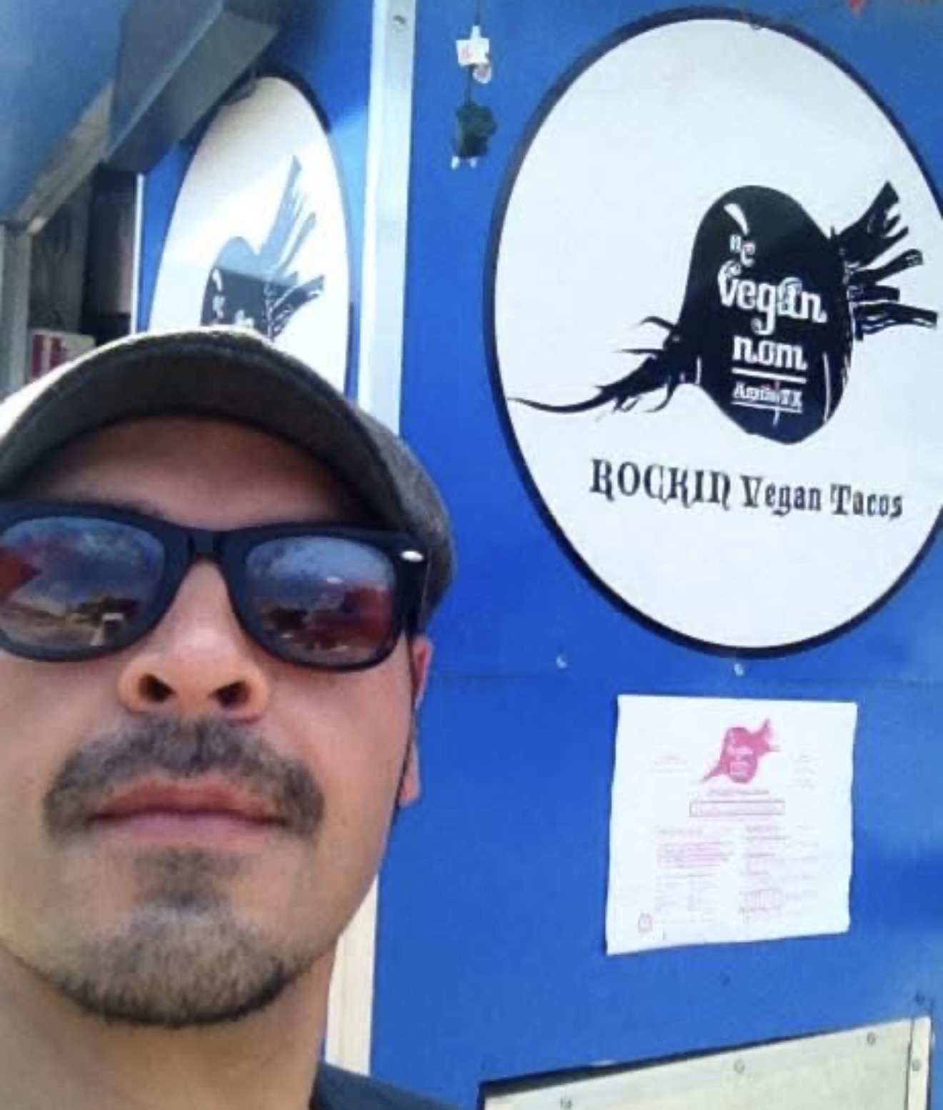 Close-up of a man wearing sunglasses and a hat, standing in front of a blue sign with a logo and text.