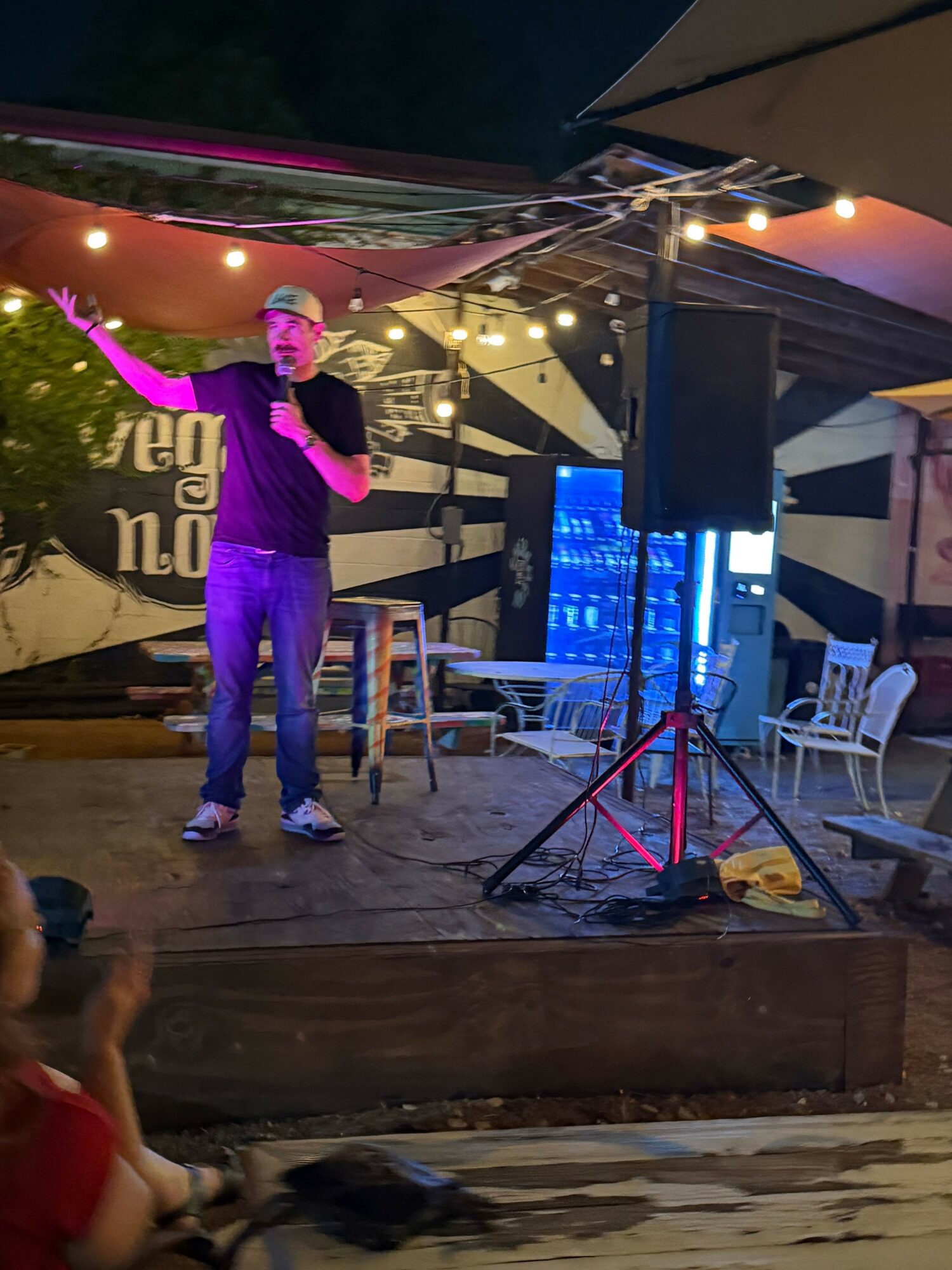 Person with pink hair and purple outfit standing on stage with equipment and chairs in background.