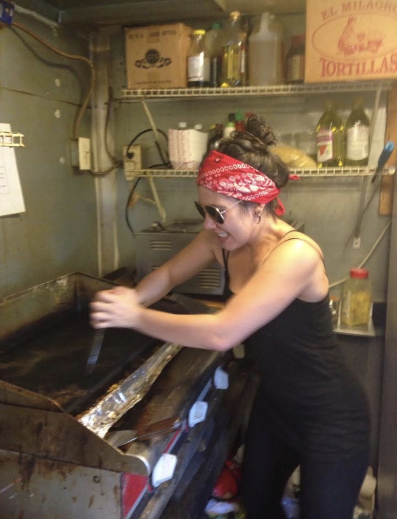 Woman wearing a red bandana and sunglasses, cooking in a small kitchen with shelves and various items.