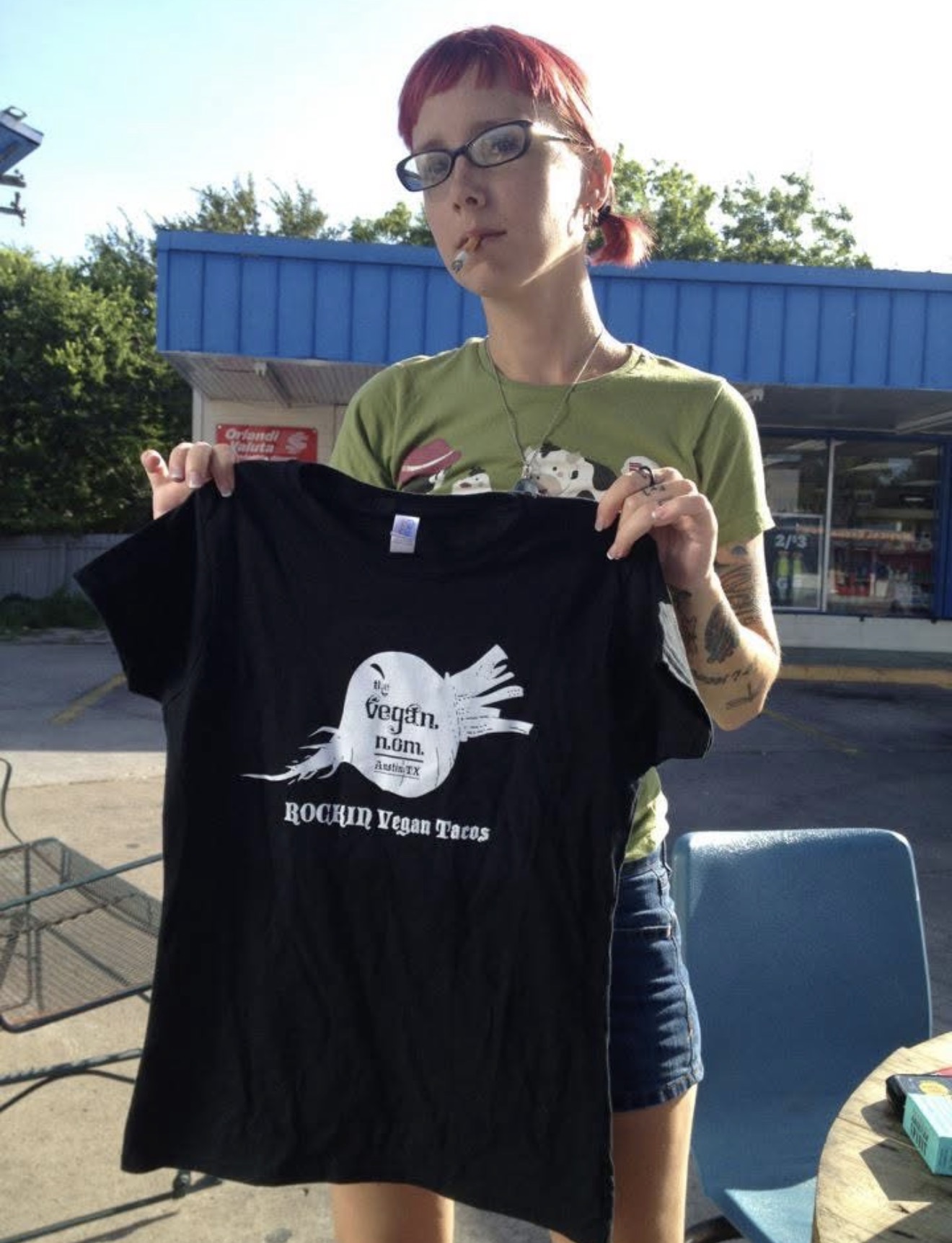 Person with red hair and glasses holding a black T-shirt outdoors, with a blue building and trees in background.