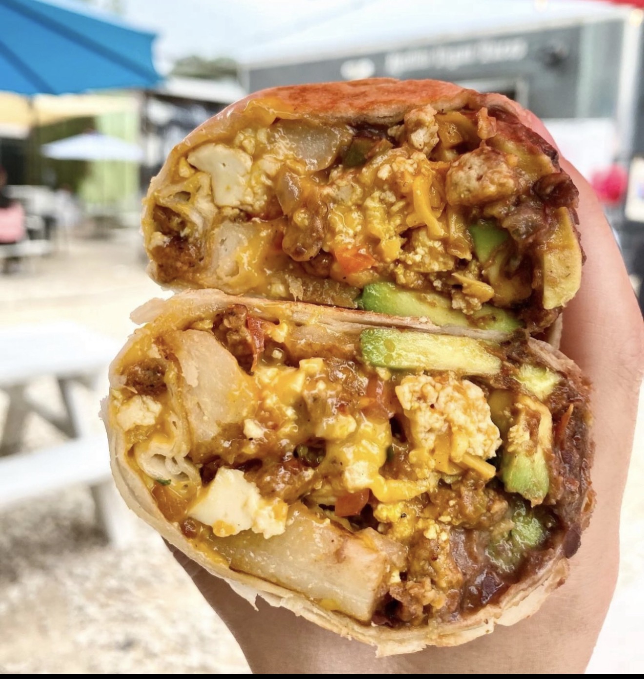 Hand holding a halved burrito with visible rice, beans, meat, and vegetables outdoors.